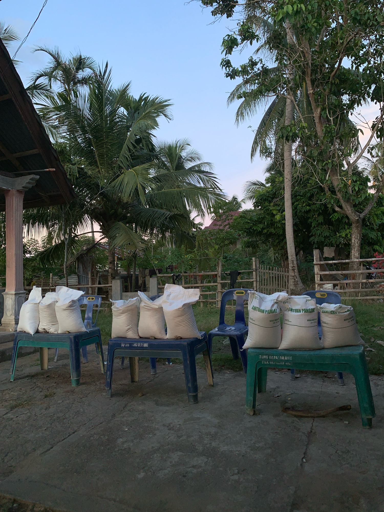 In Palohawe, Indonesia, on a gray, overcast day, eight white sacks are displayed. Four sacks rest on two green plastic tables, while the other four are placed on two blue plastic chairs. The tables are situated at the forefront, one behind the other, with the sacks on top. The chairs are positioned behind the tables, also holding sacks. The surrounding environment is rural, with a fence made of bamboo posts and branches in the background, and tall palm trees and lush vegetation dotting the landscape. A small, rudimentary structure with a corrugated metal roof is visible behind the tables and chairs. The sky above is a pale blue with hints of pink from the setting sun. The scene appears to be an outdoor gathering or distribution point.