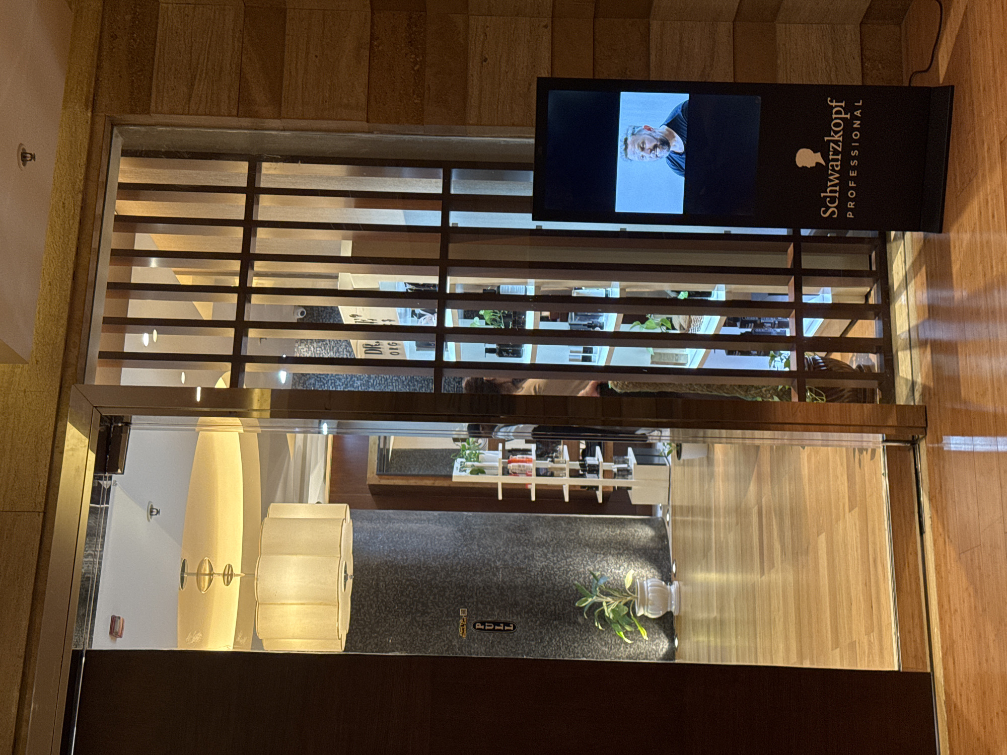 This is a full-shot, eye-level, indoor photograph of a salon entrance in Pune, India. The salon is well-lit and features polished wooden floors. A large glass door, partially open, reveals the interior. Inside, a white shelving unit displays various hair products, and a plant in a white pot adds a touch of greenery. A large, rectangular chandelier hangs from the ceiling. To the right of the entrance, a digital display screen shows a man with short, grey hair and a beard, wearing a black t-shirt. Below the man's image, the Schwarzkopf Professional logo is prominently displayed on a black stand. The exterior wall is covered in textured wooden panels. The image appears to have been taken at eye level, facing the entrance.