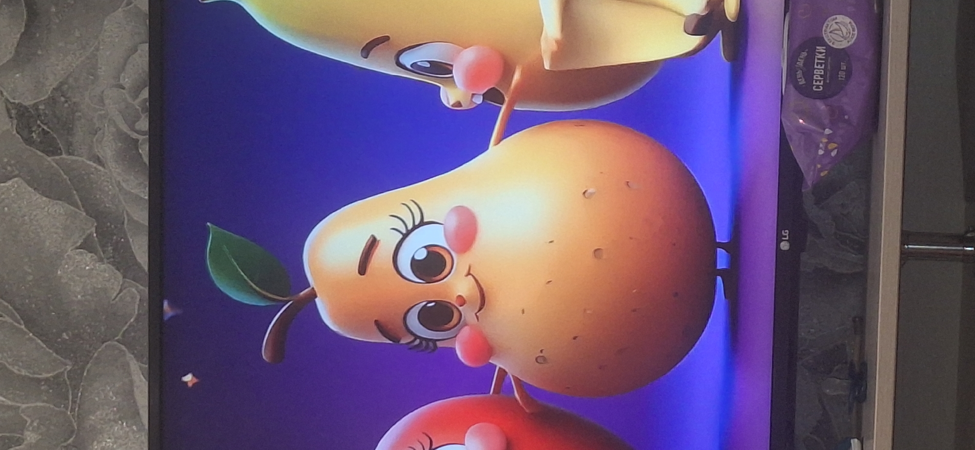 The image shows a close-up of a television screen displaying a cartoon scene with three anthropomorphic fruits. The central figure is a pear with a happy expression, rosy cheeks, large eyes with prominent eyelashes, and a stem with a leaf. To the left of the pear is a partially visible red fruit, and to the right is a yellow banana. The fruits appear to be interacting, with their arms extended towards each other. The background is a gradient of purple and blue. Below the screen, a portion of a television stand is visible, with a packet of wet wipes with Russian text on it. The image quality is moderate, with some glare from the screen. The television is likely in a home environment. No specific location in Bila Tserkva, Ukraine can be determined from the image.