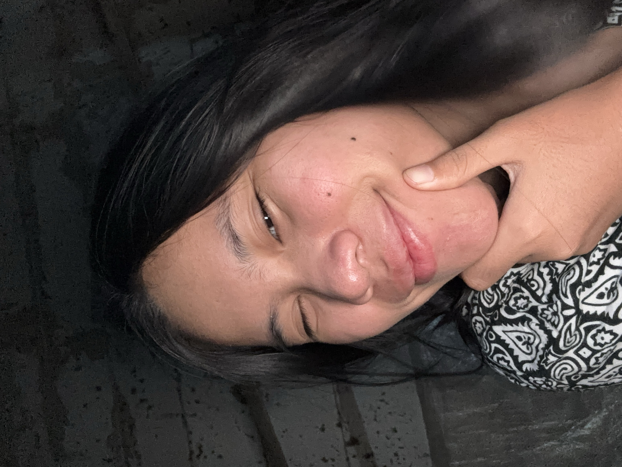 In the dimly lit interior of a building, a young woman of Asian descent is captured in a close-up selfie. Her right eye is closed in a wink, while her left eye is open, with dark hair framing her face. She has her right hand gently placed under her chin, with her fingers supporting her jawline. Her lips are slightly parted, and she appears to be smiling or smirking. She is wearing a black and white patterned shirt. The background is dark and appears to be a concrete or brick wall, with some horizontal lines suggesting shelving or structural elements. The overall impression is a casual and playful portrait taken in an indoor, possibly unfinished, space. The location is identified as East Jakarta, Indonesia.