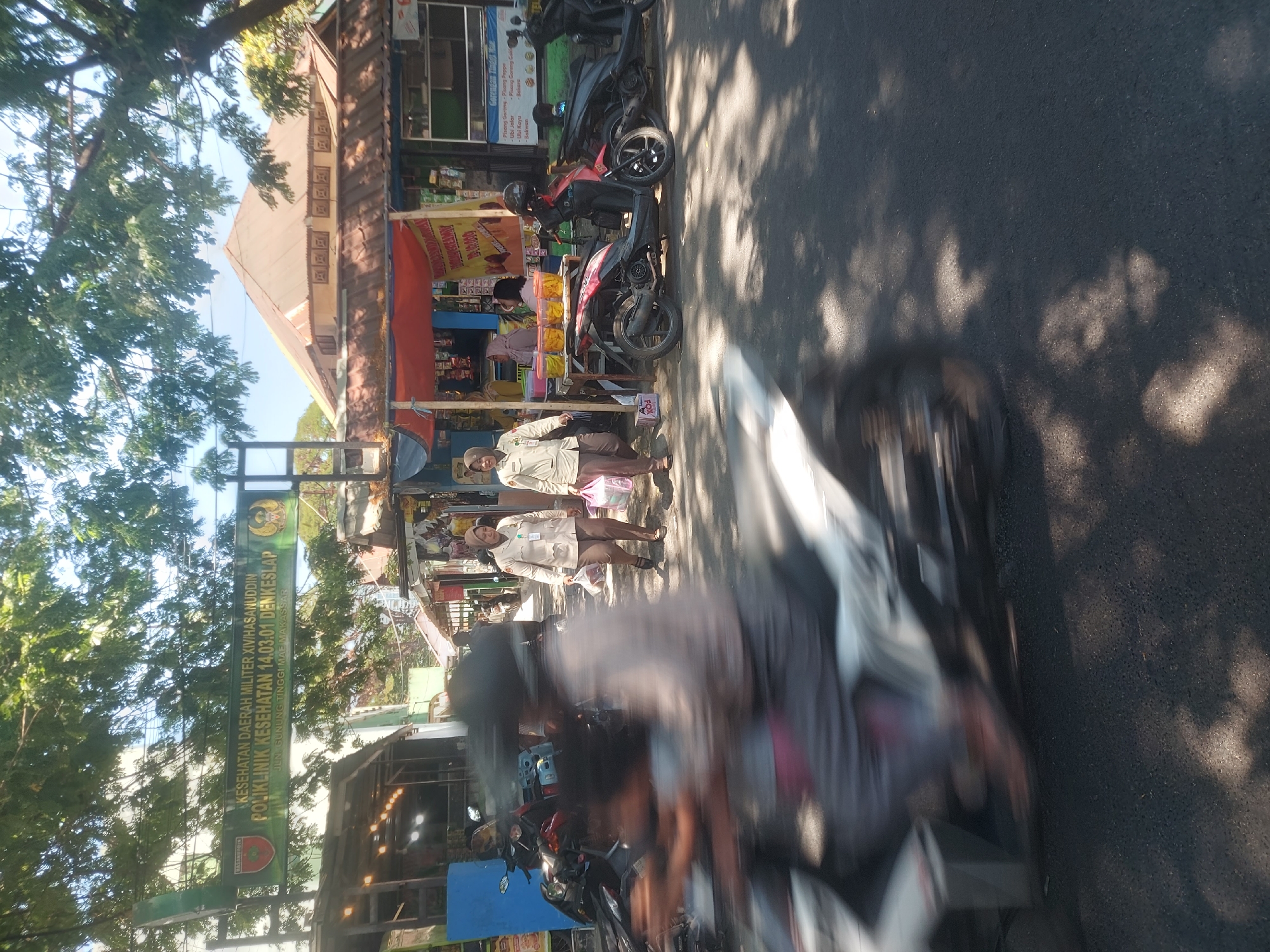 The image captures a street scene in Makassar, Indonesia, under a bright sky, with a prominent sign overhead. The sign, in Indonesian, reads "KESEHATAN DAERAH MILITER XIV/HASANUDDIN POLIKLINIK KESEHATAN 14.03.01 DENKESLAP JALAN GUNUNG TINGGI MAE MAKASSAR." This indicates the location is a military health clinic on Jalan Gunung Tinggi in Makassar. Several motorbikes are parked along the side of the road, and some are in motion, with one in the foreground blurred by motion. Two women in light-colored uniforms and headscarves are walking on the sidewalk, carrying plastic bags. Stalls are set up along the street, with one displaying various food items. Lush green trees with dappled sunlight cast shadows on the asphalt road. The overall impression is a busy urban environment with commercial activity and traffic.