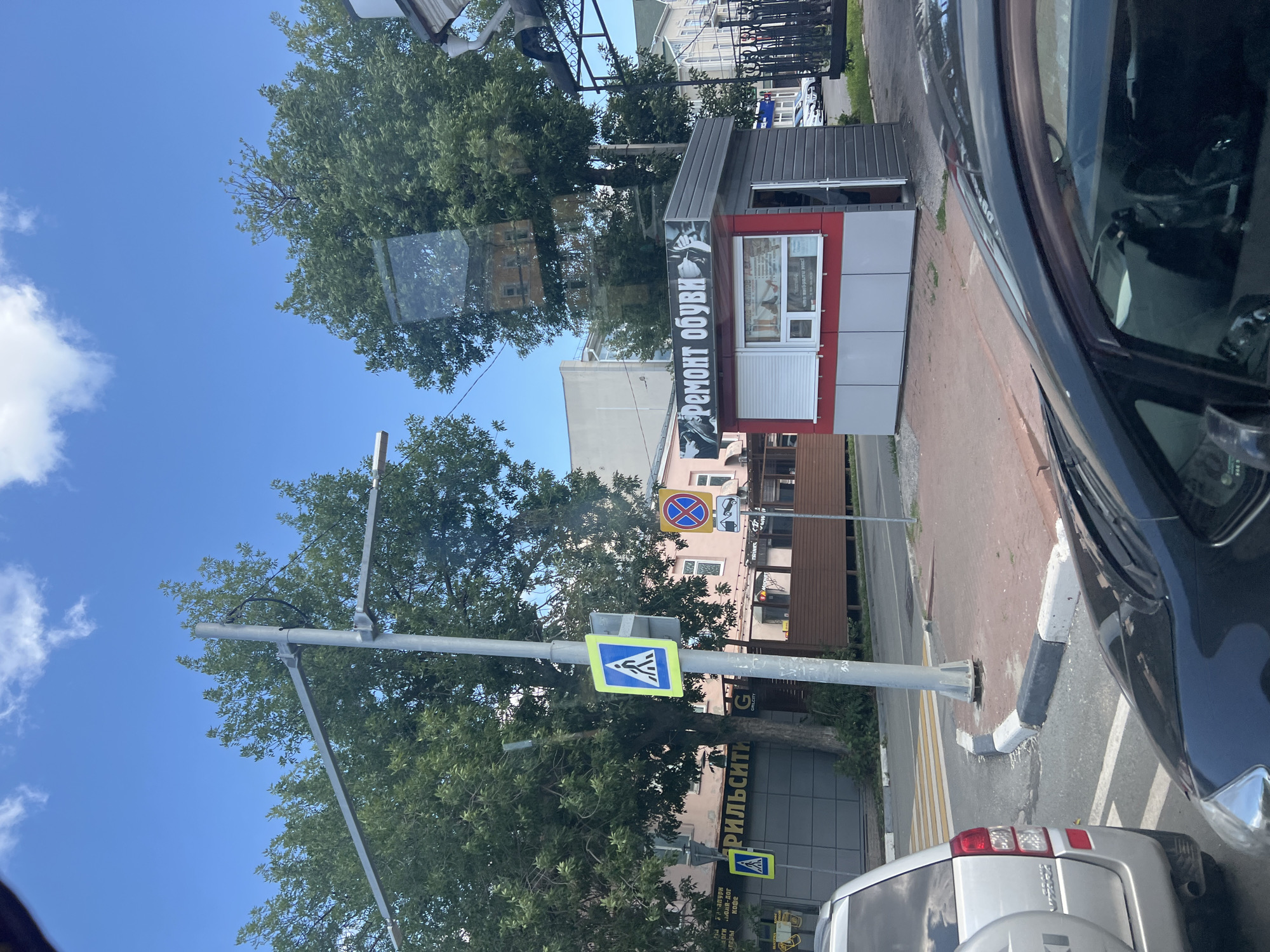 A street scene in Ulyanovsk, Russia, captures a small kiosk with a red and grey facade. The kiosk displays text in Russian that translates to "Shoe Repair." A pedestrian crossing sign is visible on a pole in front of the kiosk. The scene is observed from within a vehicle, as evidenced by reflections on the car's window and a partial view of the vehicle's interior and exterior. Trees line the street, and a blue sky with scattered clouds forms the backdrop. Other elements include road markings, a sidewalk, and a chain-link fence.