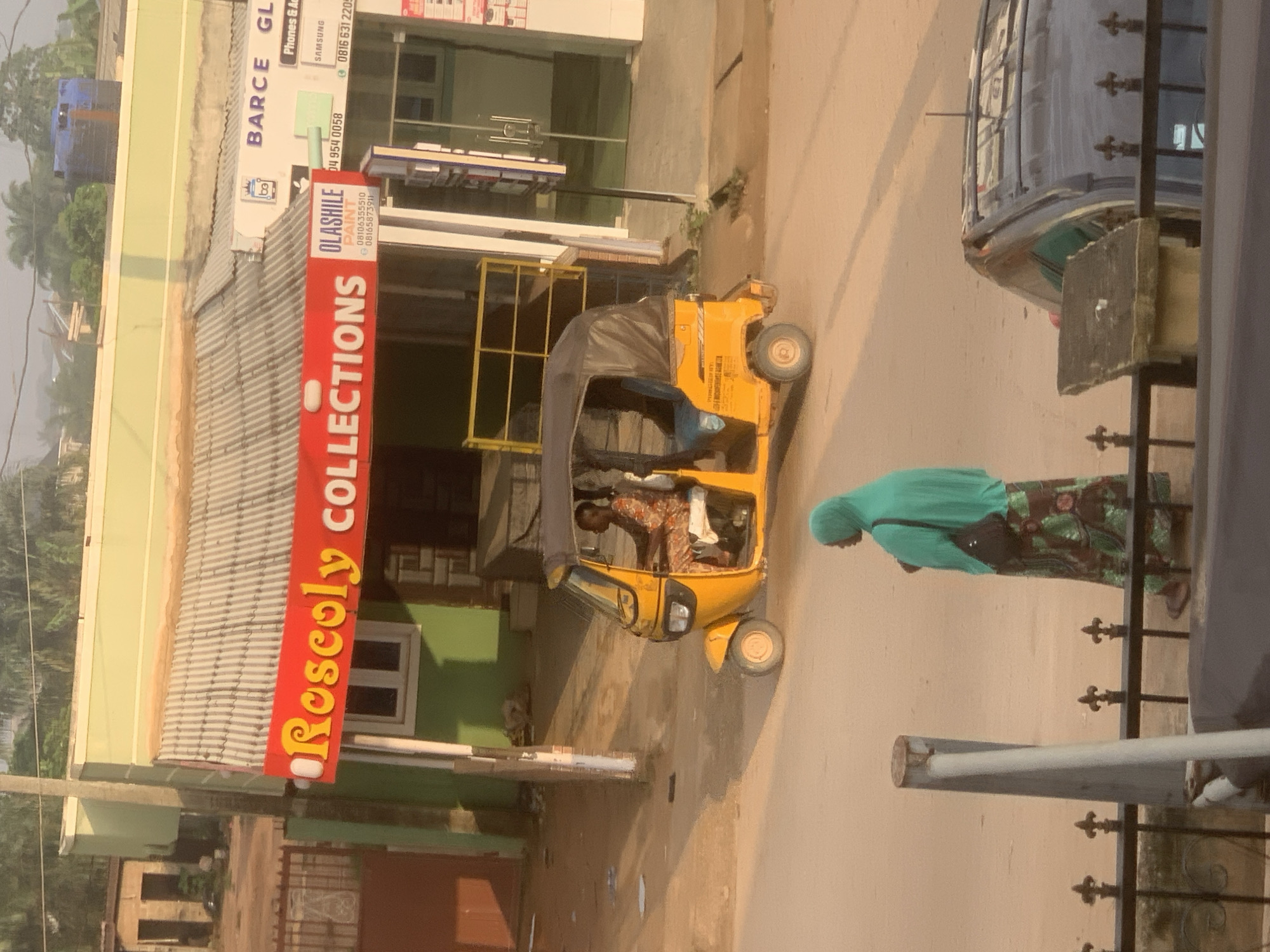 The image depicts a street scene in Shagamu, Nigeria, focusing on a small commercial building with a prominent red sign reading "Roscoly COLLECTIONS." To the right of this sign, another sign identifies "OLASHILE PAINT" with contact information. A yellow auto-rickshaw, also known as a Keke Napep, is the primary vehicle in motion, carrying two passengers. A woman wearing a teal headscarf and a patterned skirt stands on the side of the road, facing away from the camera. In the foreground, a black metal fence with decorative elements partially obscures a dark-colored car parked in the background. The environment suggests a sunny day, with harsh shadows cast on the dusty road. The buildings appear modest, typical of a commercial area in a Nigerian city.