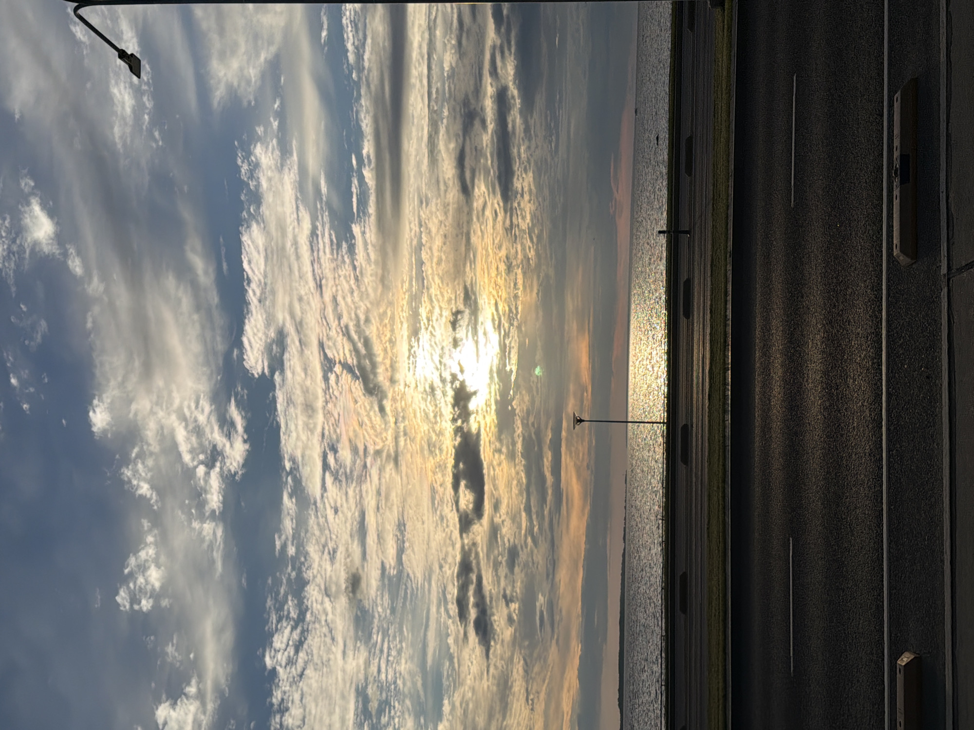 The image depicts a serene seascape at sunset, with the sun partially obscured by clouds, casting a warm glow over the water. In the foreground, a paved road runs parallel to the shoreline, with a grassy median separating it from a sidewalk and a body of water. A street lamp stands tall on the right side of the image, and another smaller lamp is visible near the water. The sky is a canvas of dramatic clouds, ranging from wispy white to darker grays, with hints of orange and yellow where the sun breaks through. The water is calm, reflecting the sky's colors, and a distant horizon line suggests a landmass or the continuation of the sea. The overall scene evokes a sense of tranquility and natural beauty, typical of a coastal setting in Tallinn, Estonia, likely along the Pirita promenade or a similar waterfront area.