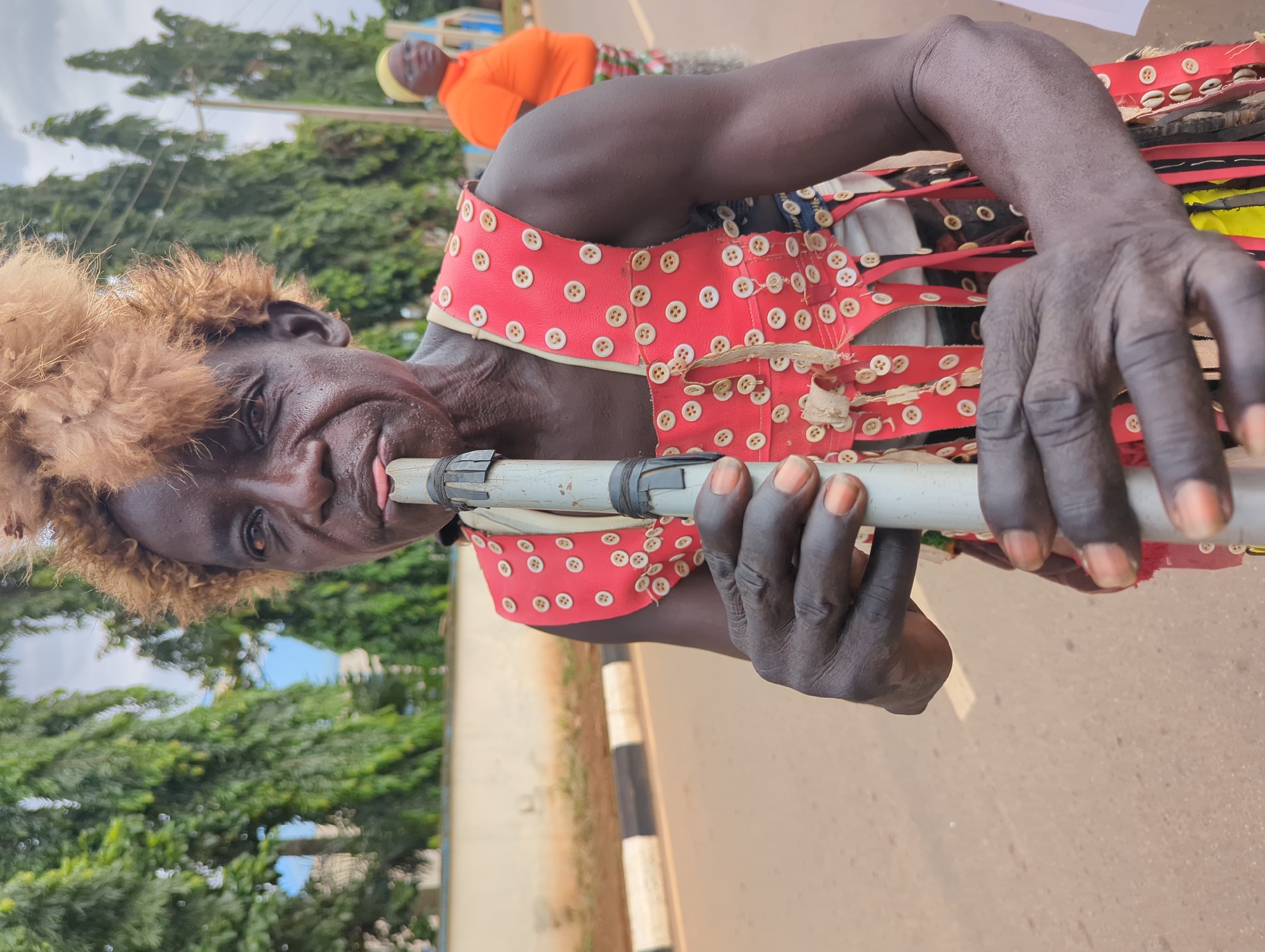 The photograph shows a man, likely an entertainer, in Maigizo, Nigeria. He is holding a white pipe, perhaps a musical instrument, to his mouth and appears to be playing it. The man is wearing a red and white costume, decorated with buttons, and a headdress made of light brown fur-like material. Another person, wearing an orange shirt and yellow head covering, can be seen in the background. The surrounding environment features green trees and a road.