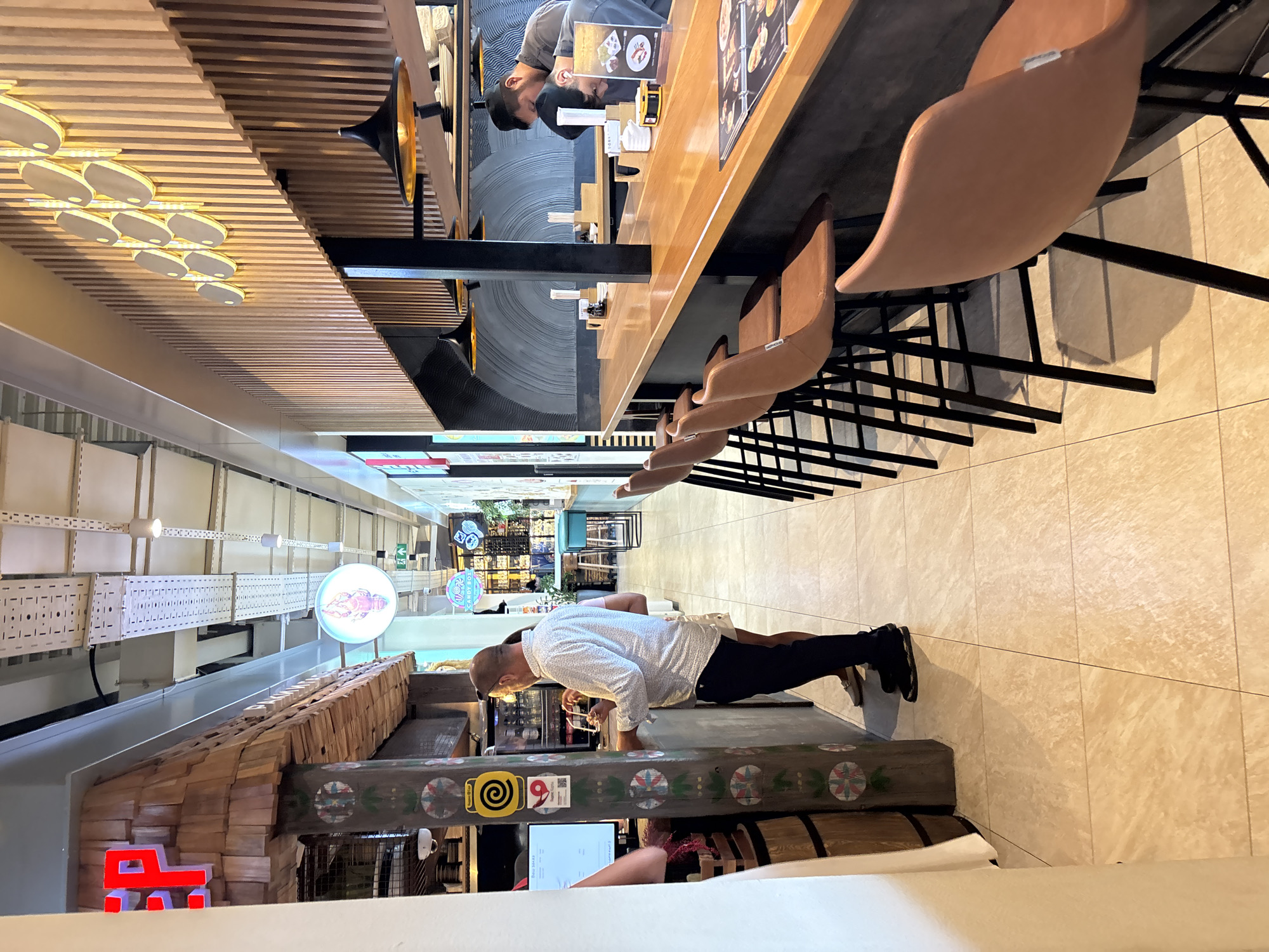 The image depicts the interior of a modern restaurant with a long wooden counter and bar stools. The counter area is well-lit, with pendant lights hanging above. The walls are decorated with vertical wooden slats and some abstract art. Several patrons are visible, some standing at the counter and others seated. In the foreground, several empty brown leather bar stools with black metal legs are lined up. The floor is tiled in a light beige color. The environment suggests a casual dining or bar setting. The image does not contain any information that would allow for a specific location within the city of Zakharovo, Russia.