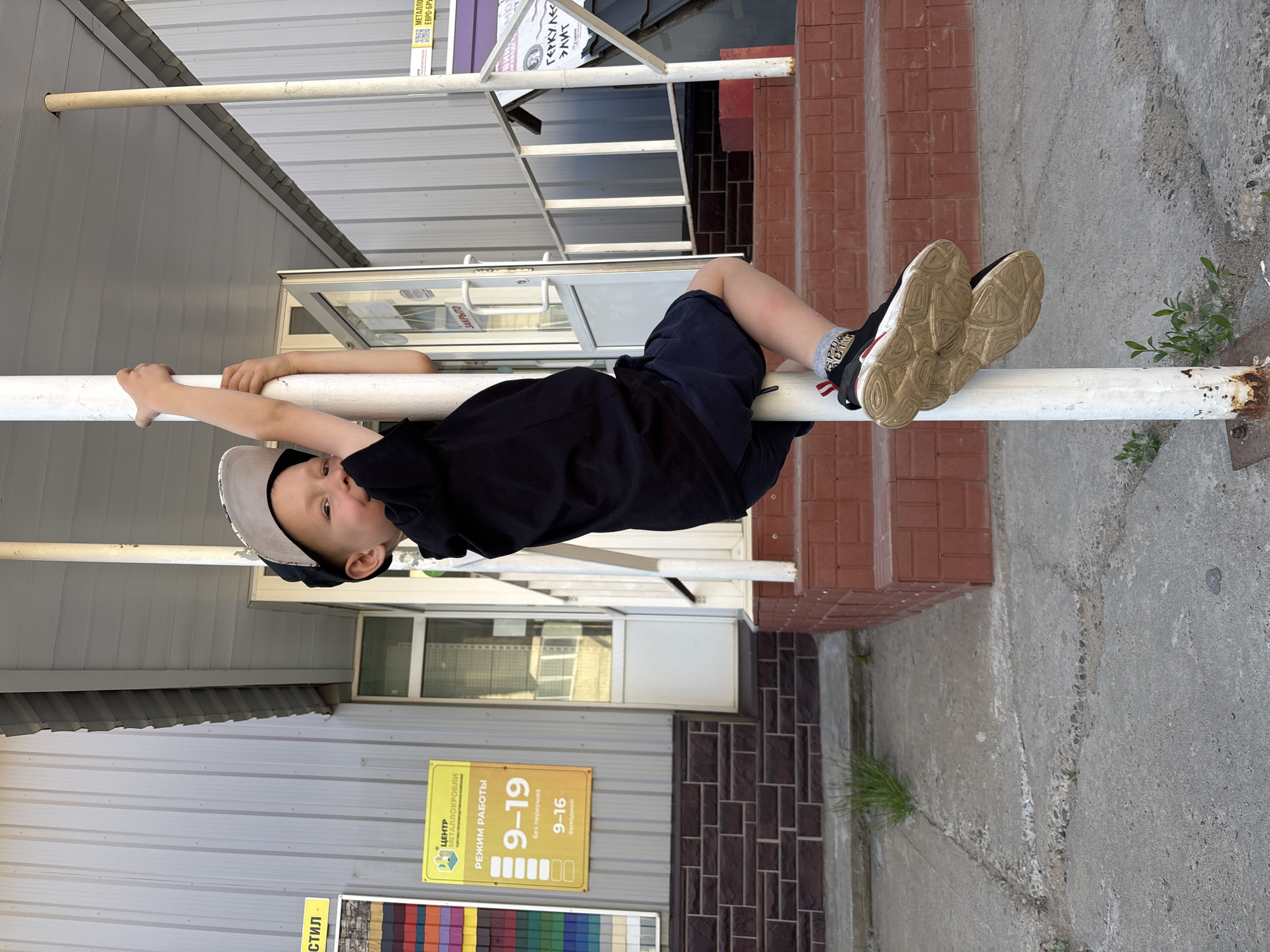 A young boy wearing a black hoodie, dark shorts, a grey and black baseball cap, and athletic shoes is climbing a white pole. He is looking at the camera and smiling. He is outdoors, in front of a building with a gray, corrugated metal exterior. There are signs on the building, one yellow sign with Russian text indicating business hours. Red brick steps lead up to a doorway. The ground is concrete with some cracks and sparse green weeds growing through. The sky is not visible in the frame. The overall impression is a candid, playful moment captured in a commercial or retail area. The location is identified as Chaikovsky, Russia, which is consistent with the visual cues of a building common in Russian commercial areas and signage in Cyrillic script.