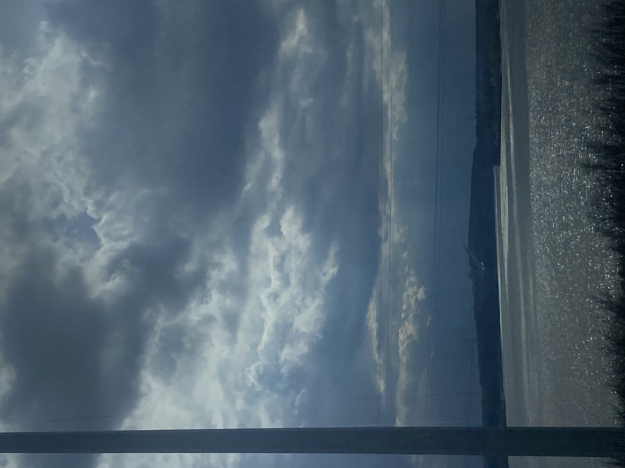 The image shows a cloudy sky above a landscape. Electrical lines are visible running across the sky. A body of water and a tree line are also visible. The picture was taken from inside a building or structure, evidenced by a visible metal beam at the bottom of the image. The location is Chaikovsky, Russia.