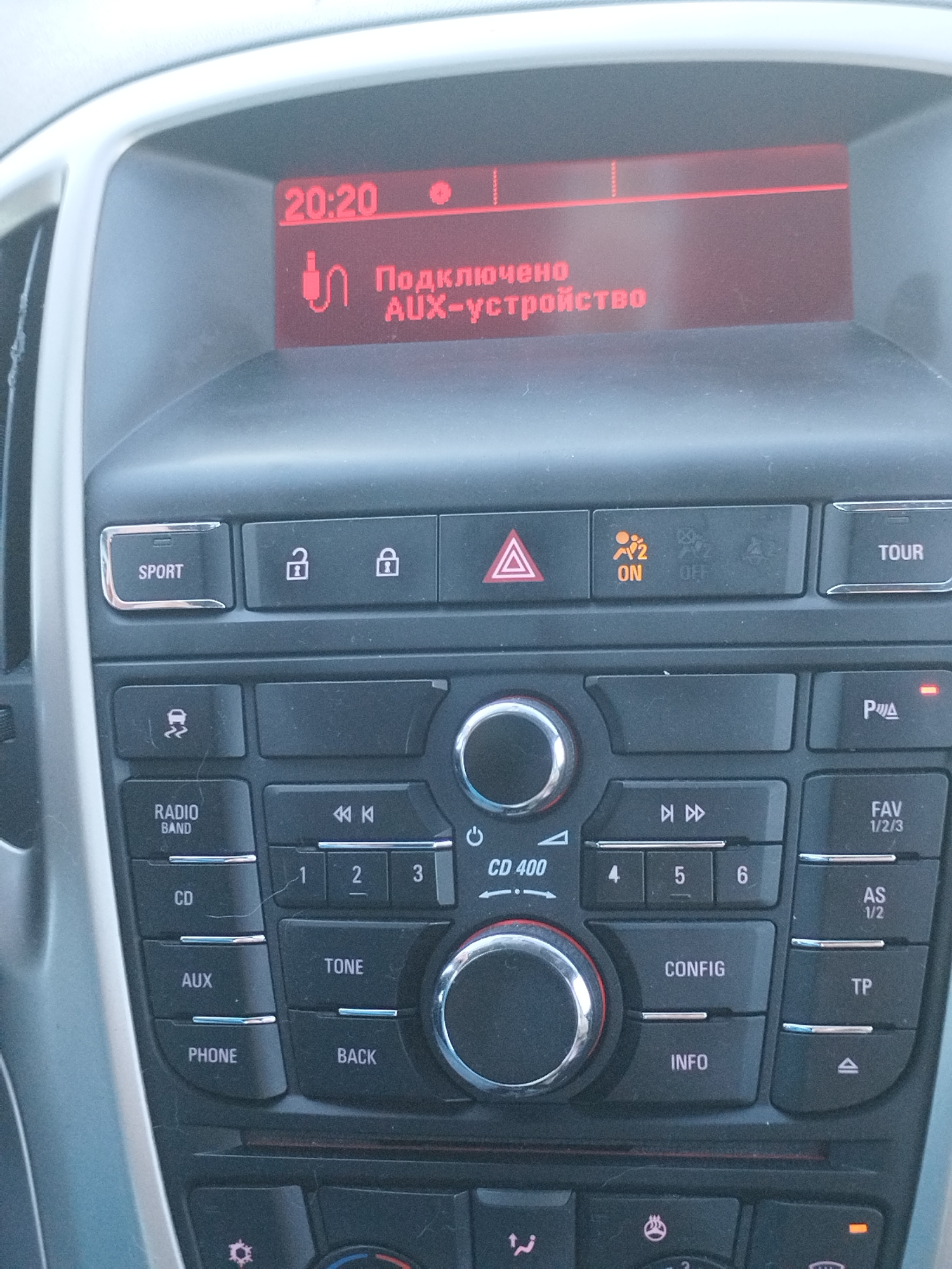 The image shows a close-up of a car's dashboard console, focusing on the infotainment system. The screen displays "20:20" at the top left and a message in Russian, "Подключено AUX-устройство" which translates to "AUX device connected," accompanied by an AUX icon. Below the screen are various buttons and controls for the car's audio system. Visible buttons include "SPORT," hazard lights, traction control, "TOUR," "RADIO BAND," "CD," "AUX," "PHONE," "CD 400," and numerical buttons for radio presets. There are also controls for playback, volume, and other functions. The console appears to be in a well-lit indoor environment, likely a car interior. There are no people or specific city locations depicted.