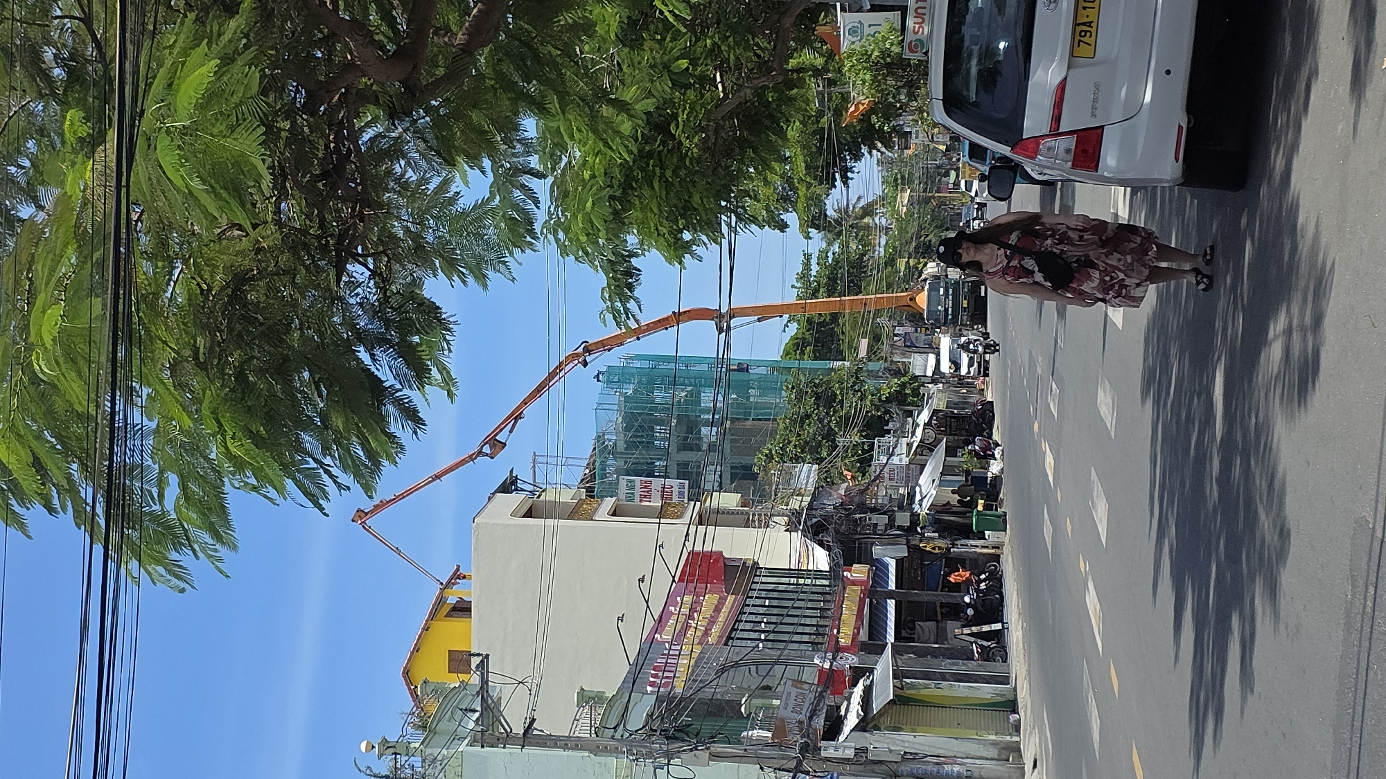 The image depicts a street scene in Nha Trang, Vietnam. A woman in a floral dress, black belt bag, sunglasses, and hat is walking on the street, next to a parked white car with license plate "79A-10". In the background, a building is under construction with green netting. An orange construction crane is positioned nearby. Overhead, power lines crisscross the sky beneath a tree canopy. Buildings with Vietnamese signage line the street. Motorcycles are parked near the shops and the road has zebra lines.