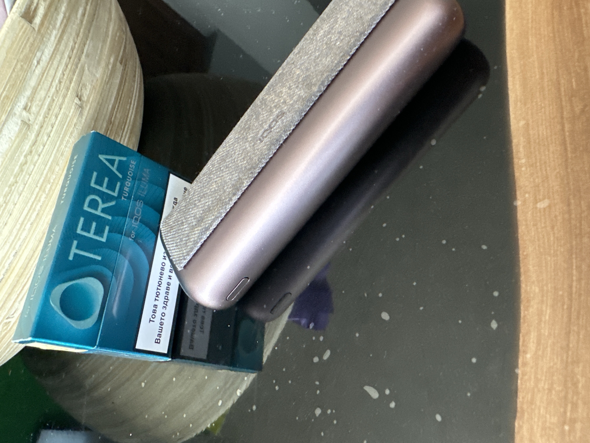 A close-up shot depicts a pack of "TEREA TURQUOISE for IQOS ILUMA" cigarettes next to an IQOS ILUMA device, which is placed on a reflective surface. The cigarette pack is light blue with the brand name "TEREA" prominently displayed in white. The text "TURQUOISE" is written in smaller font beneath the brand name. The cigarette pack also contains text in a Cyrillic script, likely Bulgarian, with the phrase "Това тютюнево из Вашето здраве и вс." The IQOS ILUMA device is cylindrical in shape with a metal casing and a fabric-covered section. The device reflects on the glass surface, creating a dark reflection. The background includes a beige object, possibly made of woven material. The scene appears to be indoors, with the focus on the cigarette pack and the electronic smoking device.