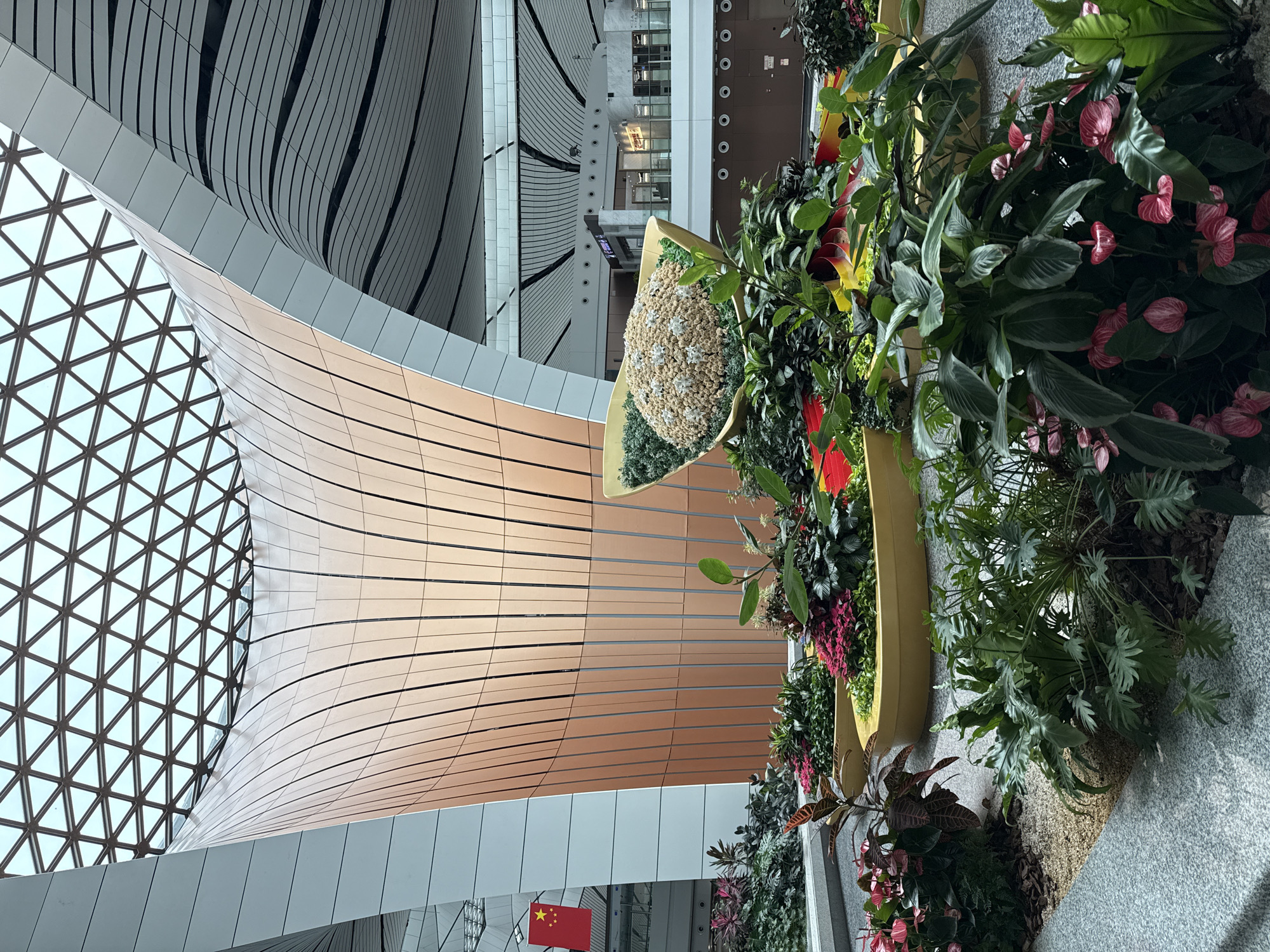 This is an interior shot of a modern, large-scale building, likely an airport terminal, with a distinctly organic architectural design. The scene is dominated by a massive, curved structure clad in vertically aligned copper-toned panels, which culminates in a broad opening at the top, revealing a geometric glass ceiling. The interior space is vast, with multiple levels visible in the background, featuring sleek white walls and a grid-like ceiling structure.
In the foreground, a vibrant and lush botanical display is arranged in several large, elongated, yellow planters. These planters contain a diverse collection of foliage and flowers, including deep green, broad-leafed plants, plants with striking red and pink leaves, and clusters of bright pink anthurium flowers. One planter holds a large, mound-like floral arrangement, seemingly made of small beige and white elements, perhaps dried flowers or decorative stones, accented with delicate white flower-like embellishments. The ground surrounding the planters is covered in a mix of small gravel and wood chips.
A Chinese flag, red with yellow stars, is visible hanging on the left side of the frame, attached to a white pillar, suggesting the location is within China. The overall impression is one of spaciousness, contemporary design, and a deliberate integration of nature within an artificial, high-tech environment. The location is identified as Guangyang District, China, which is consistent with the presence of a major international airport or similar large public infrastructure. There are no people visible in the image.