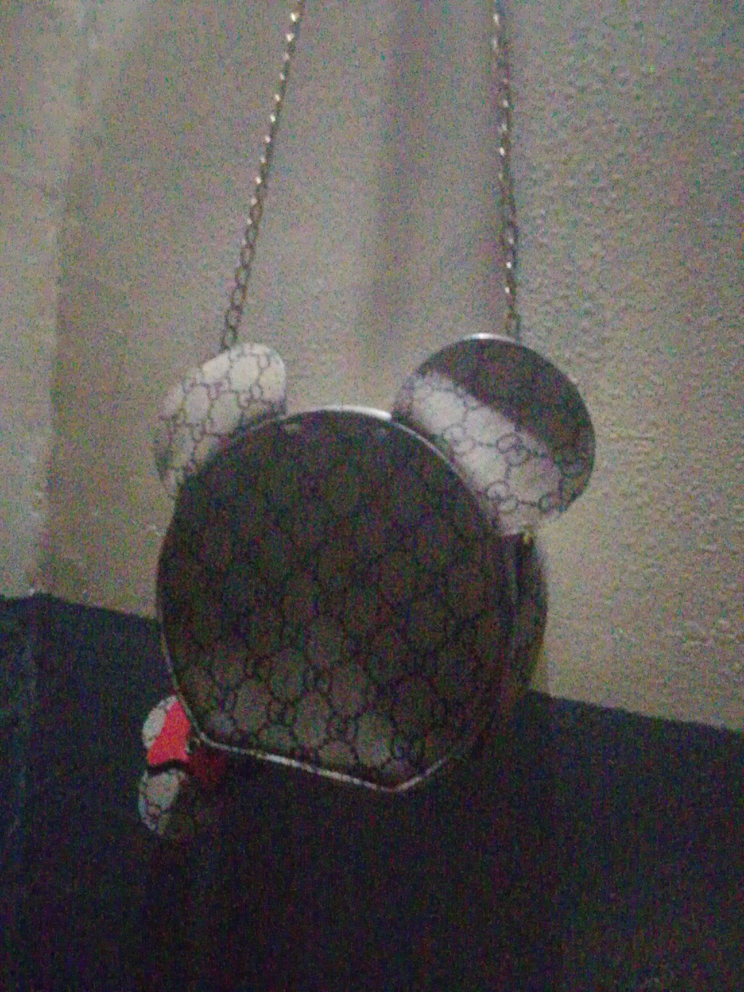 A low-resolution, grainy image depicts a single handbag suspended against a textured wall. The handbag is designed in the shape of a stylized cartoon character's head, featuring a large circular main body and two smaller circular elements resembling ears attached to the top. The visible surfaces of the bag display a repeating, interlocking geometric pattern in a light color (possibly beige or off-white) with darker grey or brown designs. A thin, metallic chain strap is attached to the bag and extends upwards, out of frame. A small, partially visible element, appearing to be light grey with a red section, is present at the bottom-left edge of the main bag body. The background consists of a uniformly textured, light-colored wall surface. A darker, horizontal surface is partially visible at the bottom edge of the frame. The scene is illuminated by dim ambient light, resulting in significant image noise. The image provides no specific location information within the city of Unknown, Unknown beyond an indoor setting.