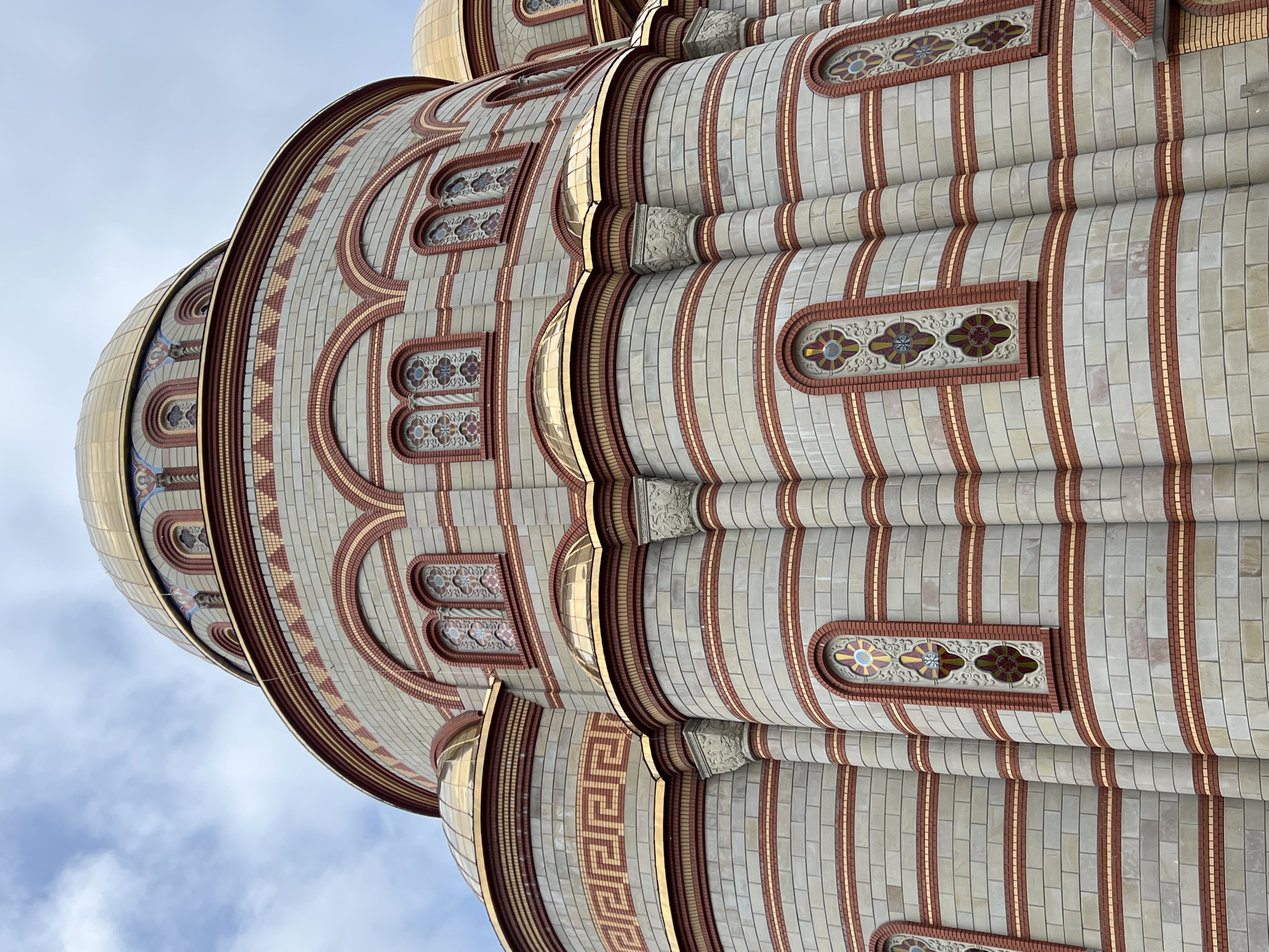 A close-up, low-angle view captures the ornate exterior of a large Eastern Orthodox cathedral, likely the Cathedral of Saints Volodymyr and Olha in Ternopil, Ukraine. The image highlights the intricate architectural details of the building's upper sections and domes against a soft, cloudy blue sky.
The cathedral's facade is primarily clad in light grey or beige rectangular tiles, arranged in vertical bands. These bands are separated by vertical stripes of reddish-brown brickwork and slender rows of small golden or yellow accent tiles, creating a visually rich, rhythmic pattern. Recessed, arched window-like openings punctuate the tiled surfaces, framed by reddish-brown bricks. Within these arches are decorative panels, resembling stained glass or mosaics, featuring elaborate floral and geometric designs in shades of red, blue, green, and yellow.
Multiple golden domes are visible, some partially, reflecting the daylight. Below the domes, drum sections are adorned with similar tiling patterns. One section distinctively features a repeating Greek key motif rendered in reddish-brown bricks and light-colored tiles. Additionally, carved stone elements, possibly capitals or corbels, depicting intricate foliage patterns, are visible supporting structural features. The overall impression is one of grandeur and meticulous craftsmanship. There are no people or discernible activities in the image, allowing the architectural beauty to be the sole focus.