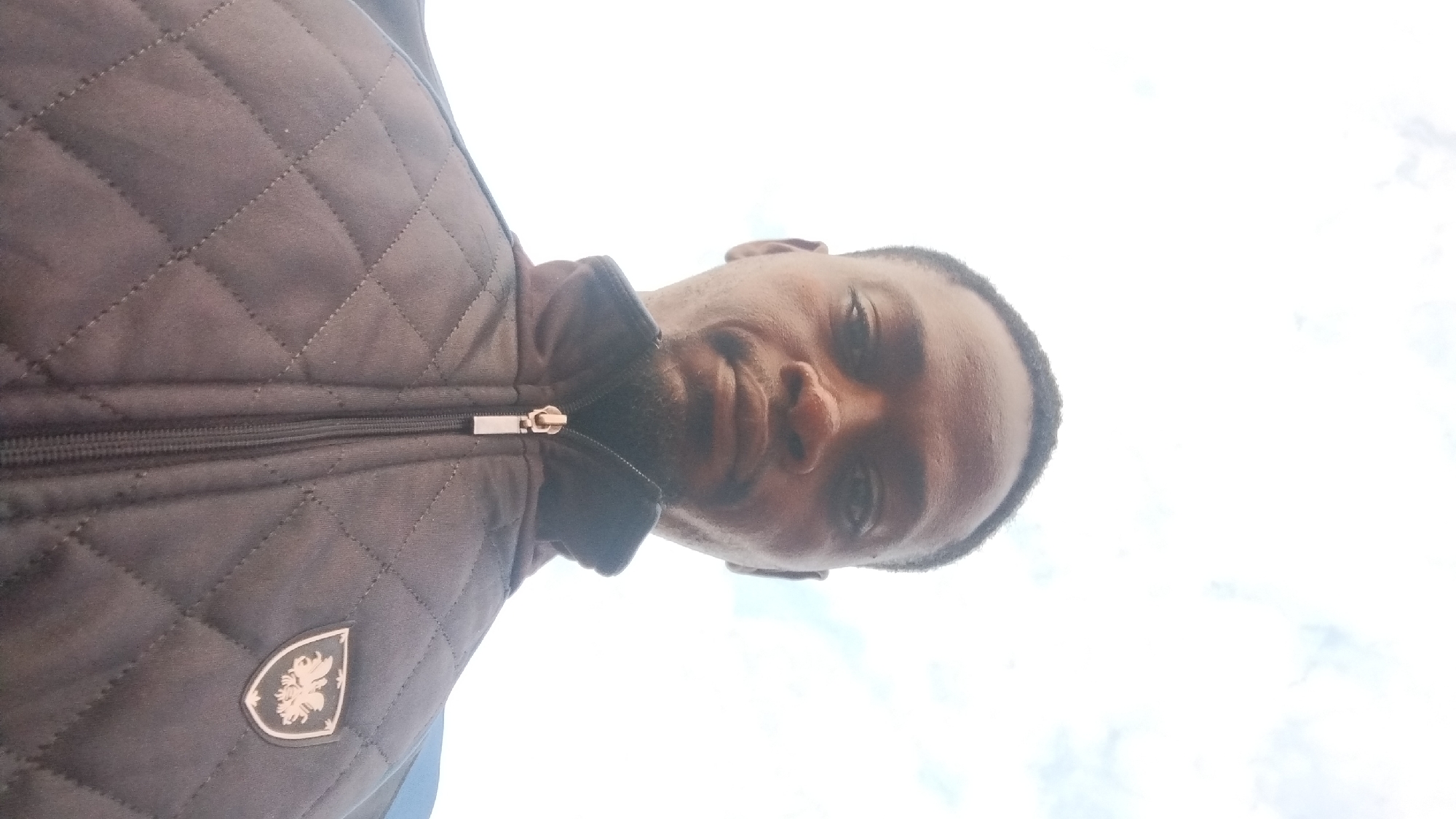 The media file is a selfie taken from a low angle, looking up at a man. The man is wearing a dark, quilted jacket with a zipper. A silver zipper pull is visible. On the left side of the jacket, there is a shield-shaped emblem with a crest inside. The man has dark skin and short, dark hair. He is looking towards the camera with a neutral expression. The background is a bright, overcast sky with some clouds. The lighting suggests it is daytime, but the overcast conditions make it difficult to determine the exact time. There are no visible location cues in the image. The image appears to be taken outdoors.