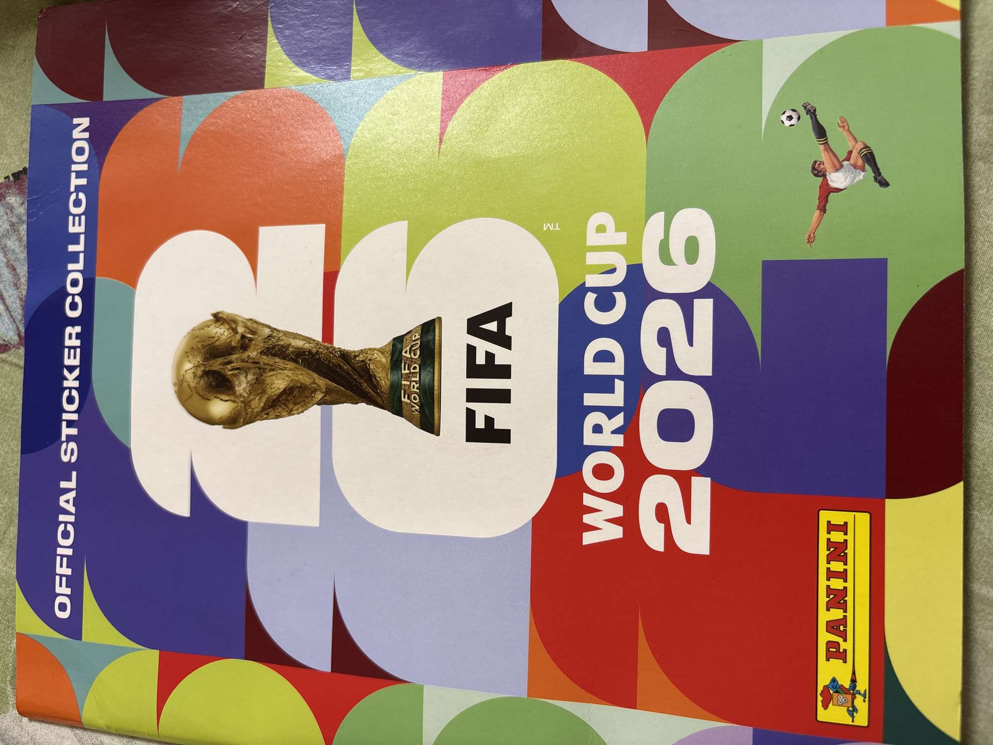 The image shows the cover of an official sticker collection for the FIFA World Cup 2026. The cover features the FIFA World Cup trophy, the text "OFFICIAL STICKER COLLECTION", "FIFA WORLD CUP 2026", and the "PANINI" logo. A stylized illustration of a soccer player kicking a ball is also present. The cover is designed with a colorful, abstract pattern of geometric shapes and overlapping circles in various hues of purple, blue, green, orange, red, and maroon. The image does not provide any information or visual cues that would allow for the determination of a location in the city of Ветлужский, Russia. The scene is a close-up shot of the sticker collection cover, likely taken indoors.
