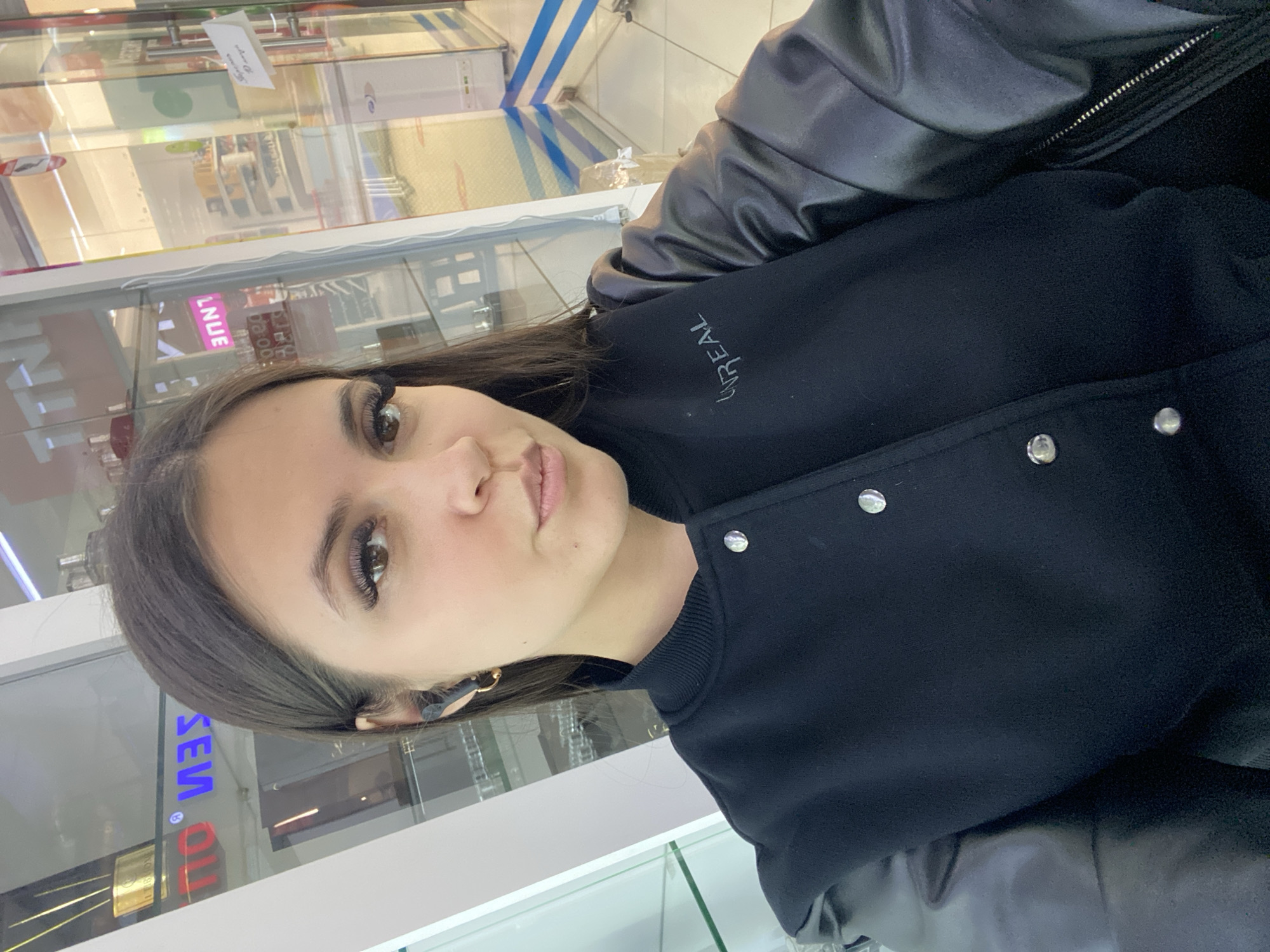 A young woman with dark hair and brown eyes is featured in a close-up, selfie-style photograph, taken indoors in Chaikovsky, Russia. She looks directly at the camera with a neutral expression, showcasing prominent long eyelashes. She wears a black jacket with silver buttons and a contrasting shiny material, possibly faux leather, on the shoulders or collar. The "L'ORÉAL" logo is visible stitched in white on the jacket's chest. An earring and a black earbud or hearing aid are present in her visible ear. The background is out of focus, revealing reflections from what appear to be glass display cases or windows. Visible reversed text in the reflections includes "L NUE," "OLLI," and "SEV," along with numerical digits like "00" and "20." A small white paper label with indistinct text is also reflected. The setting suggests a commercial or retail space, illuminated by artificial lighting.