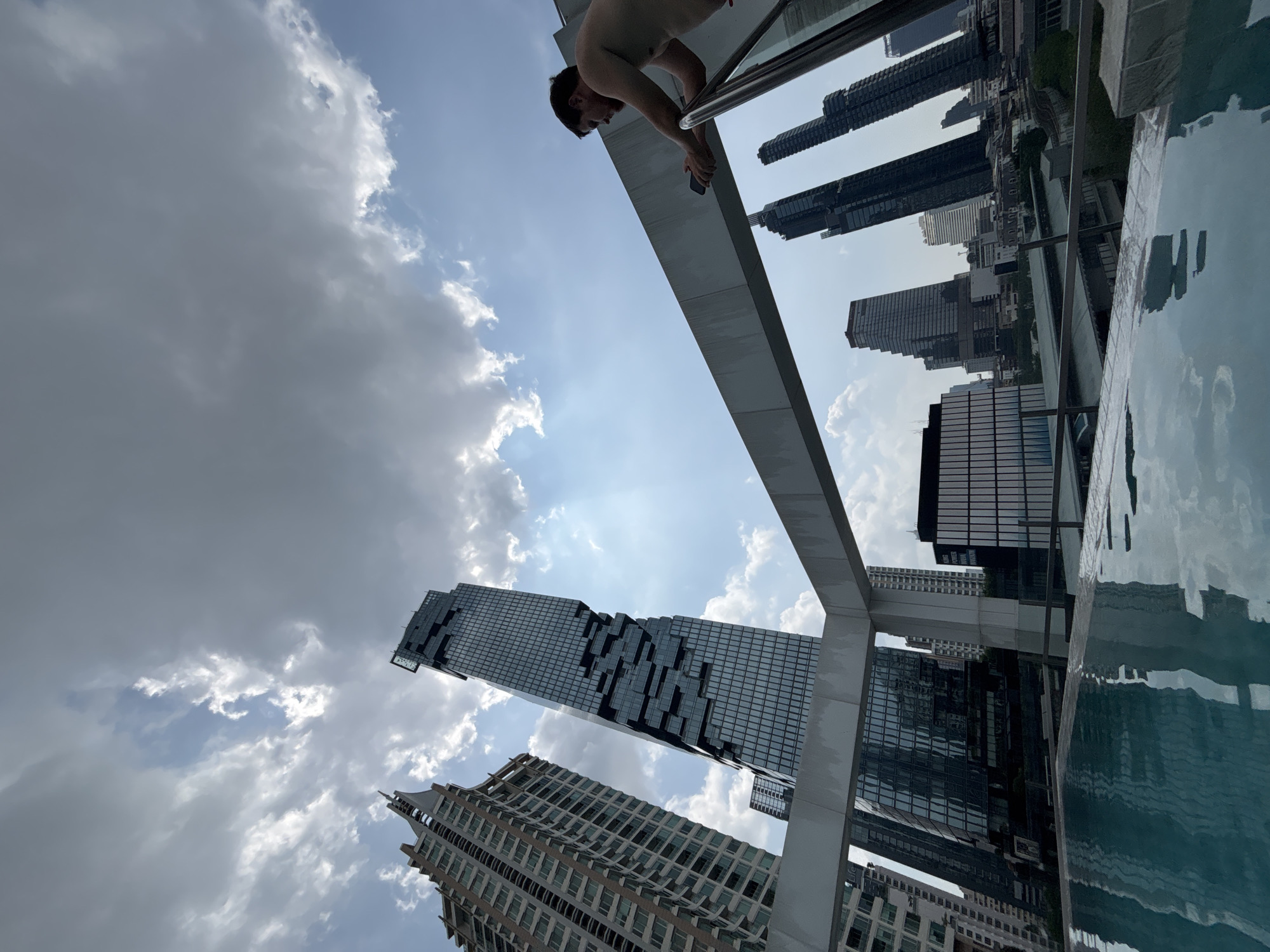 This image, captured from a low, upward-looking perspective and rotated counter-clockwise, presents a scene from a high-rise in Bangkok, Thailand. A shirtless man is prominently featured, leaning over a metal railing, seemingly gazing downwards or using a smartphone he holds in his hand.
The backdrop is dominated by a dynamic urban skyline under a partly cloudy sky. The iconic MahaNakhon Tower, with its distinctive "pixelated" architectural design, stands out as a central focus among several other skyscrapers. Sunlight streams through gaps in the clouds, creating bright rays across the blue sky.
To the right of the frame, the calm, reflective surface of a swimming pool mirrors the surrounding buildings, including glimpses of modern glass facades. The structural elements of the building, such as a sturdy concrete beam and the railing the man holds, frame the view, emphasizing the elevated position. The time of day is clearly daytime, marked by the ample natural light. The varying cloud formations range from soft white to more opaque grey. There is no visible text in the image. The composition highlights the grandeur of modern urban architecture and a unique high-altitude leisure setting in the heart of the city.