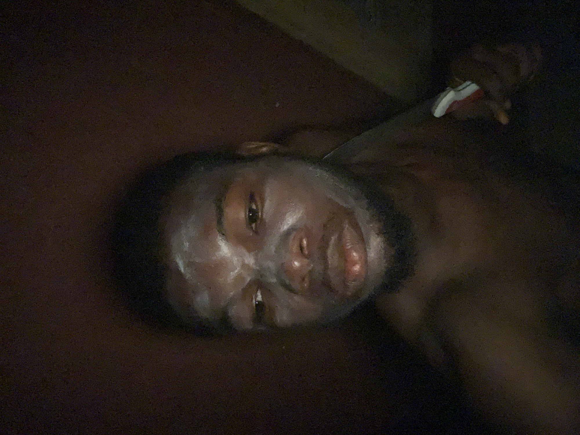 The image shows a man lying on a dark surface in a poorly lit environment. His face has a white substance applied to it. A hand holding a white and red object is visible near his shoulder. The location is unknown, but the prompt suggests Onitsha, Nigeria. Specific details about the man's condition, the substance on his face, the object in the hand, and the nature of the environment are unclear due to the image quality and limited context.
