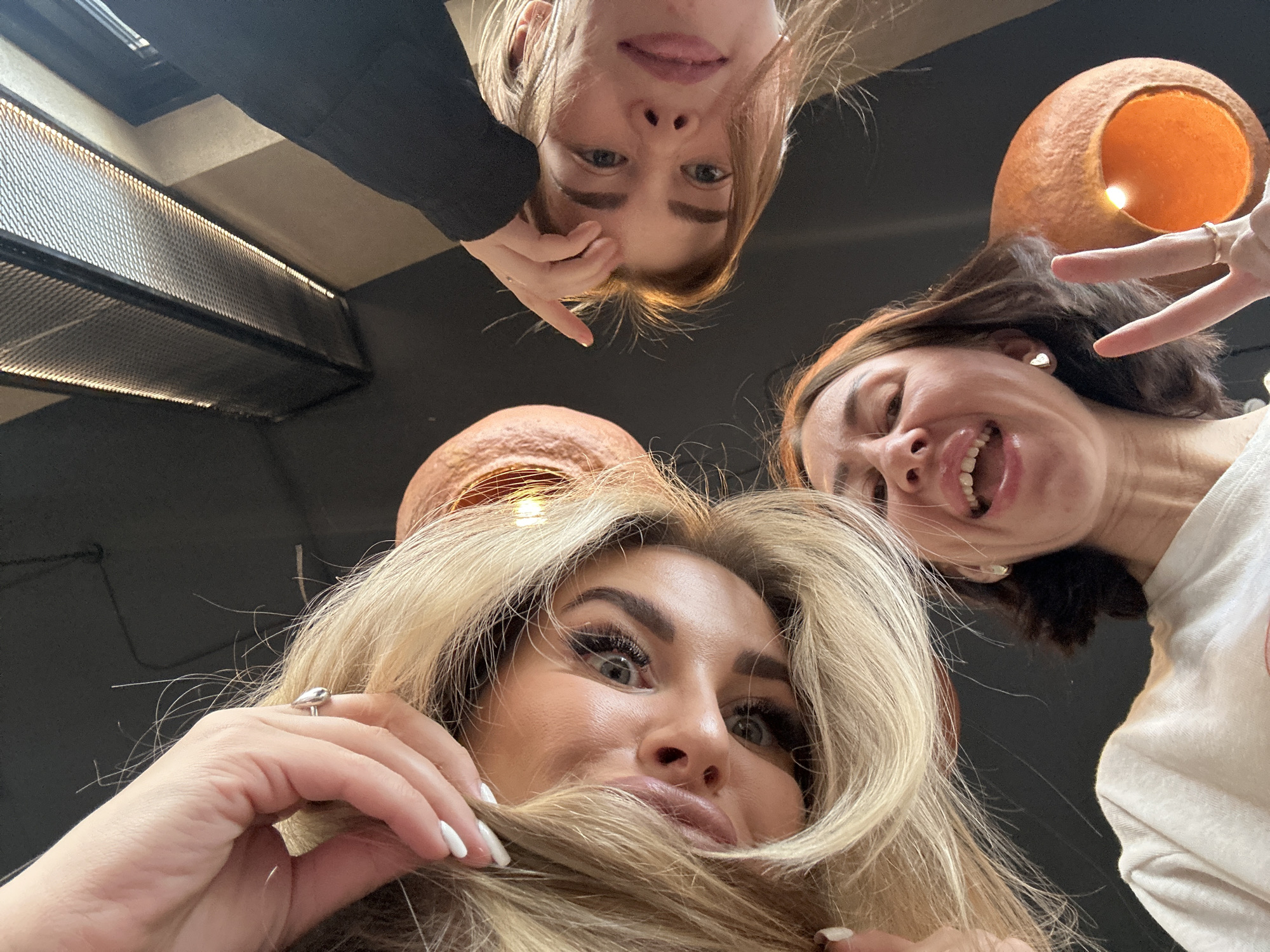 An indoor image taken from a low angle in Усады, Russia, depicts three women looking down directly into the camera. The woman on the left, with long blonde hair, is looking directly forward, showing prominent eye makeup, and her left hand with white nail polish and a silver teardrop ring is raised near her hair. The woman at the bottom center, with shorter brown hair, is laughing with her mouth open and making a peace sign with her right hand, which bears a gold-toned ring. She also wears a heart-shaped earring. The woman on the right, with lighter brown hair, is looking downwards, with her left hand touching her forehead. The background is dark, featuring two prominent bell-shaped terracotta light fixtures emitting an orange glow, and a long, rectangular industrial light fixture with a metallic mesh grille visible in the upper right.