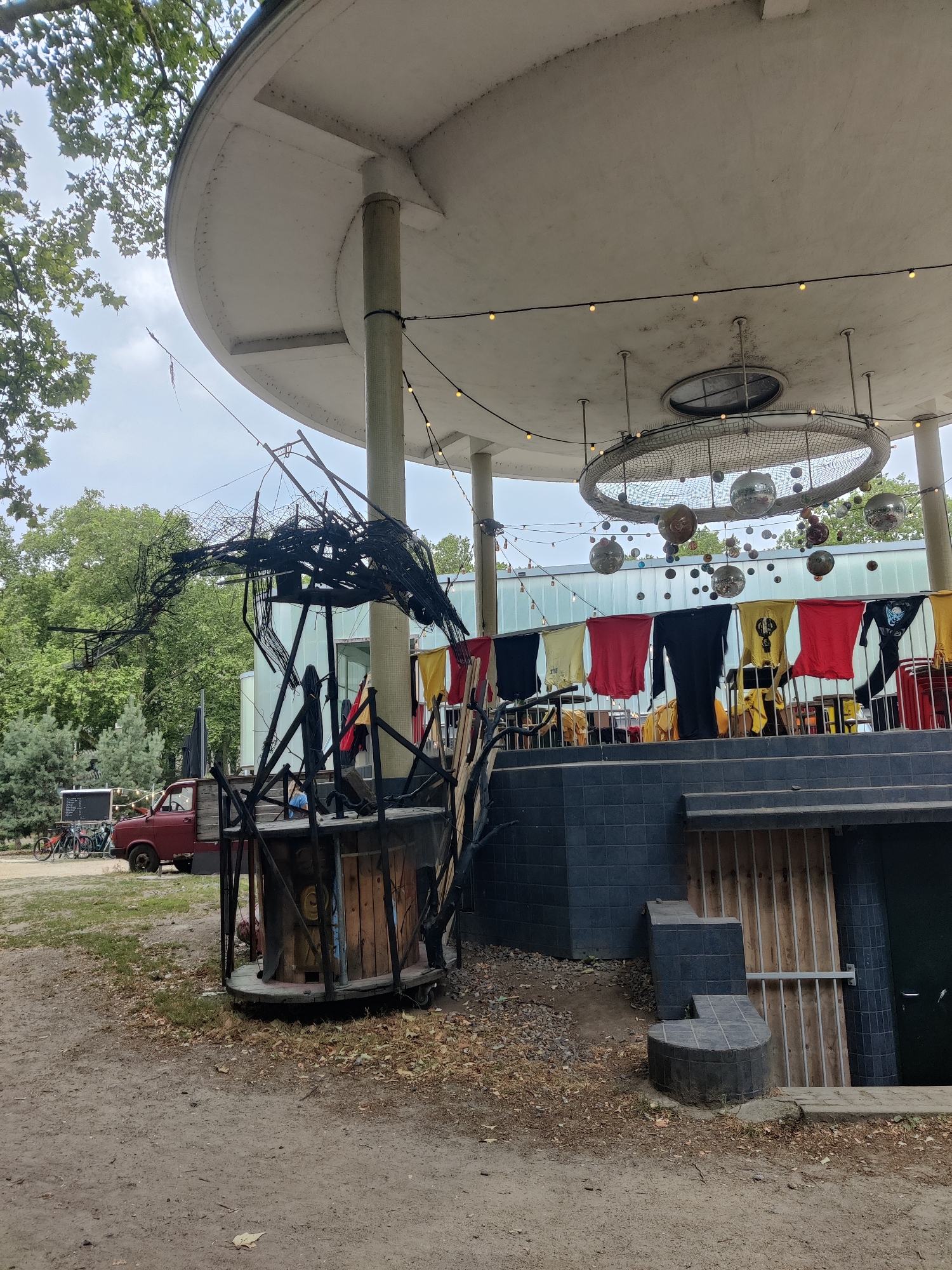 The image shows an outdoor area with a circular, white-roofed structure supported by pillars, reminiscent of a pavilion or stage. The structure has a distinctive, modern design. Suspended from the ceiling of the structure are strings of yellow lights and a large, circular net with various reflective spheres hanging from it. Below the roofline, a row of brightly colored t-shirts or flags are displayed, hanging from a railing.
In the foreground, there is a large, black, sculptural installation made of metal and wood, resembling a stylized bird or creature in flight. It is positioned in front of the pavilion. To the left of the sculpture is a red truck and a row of bicycles parked on a dirt path.
The ground is covered with dirt and fallen leaves, indicating an outdoor, perhaps park-like setting. Behind the pavilion, there are glass windows and trees, suggesting it is situated within a larger green space. The overall atmosphere appears to be casual and possibly related to an event or market.
The image was taken in Liège, Belgium.