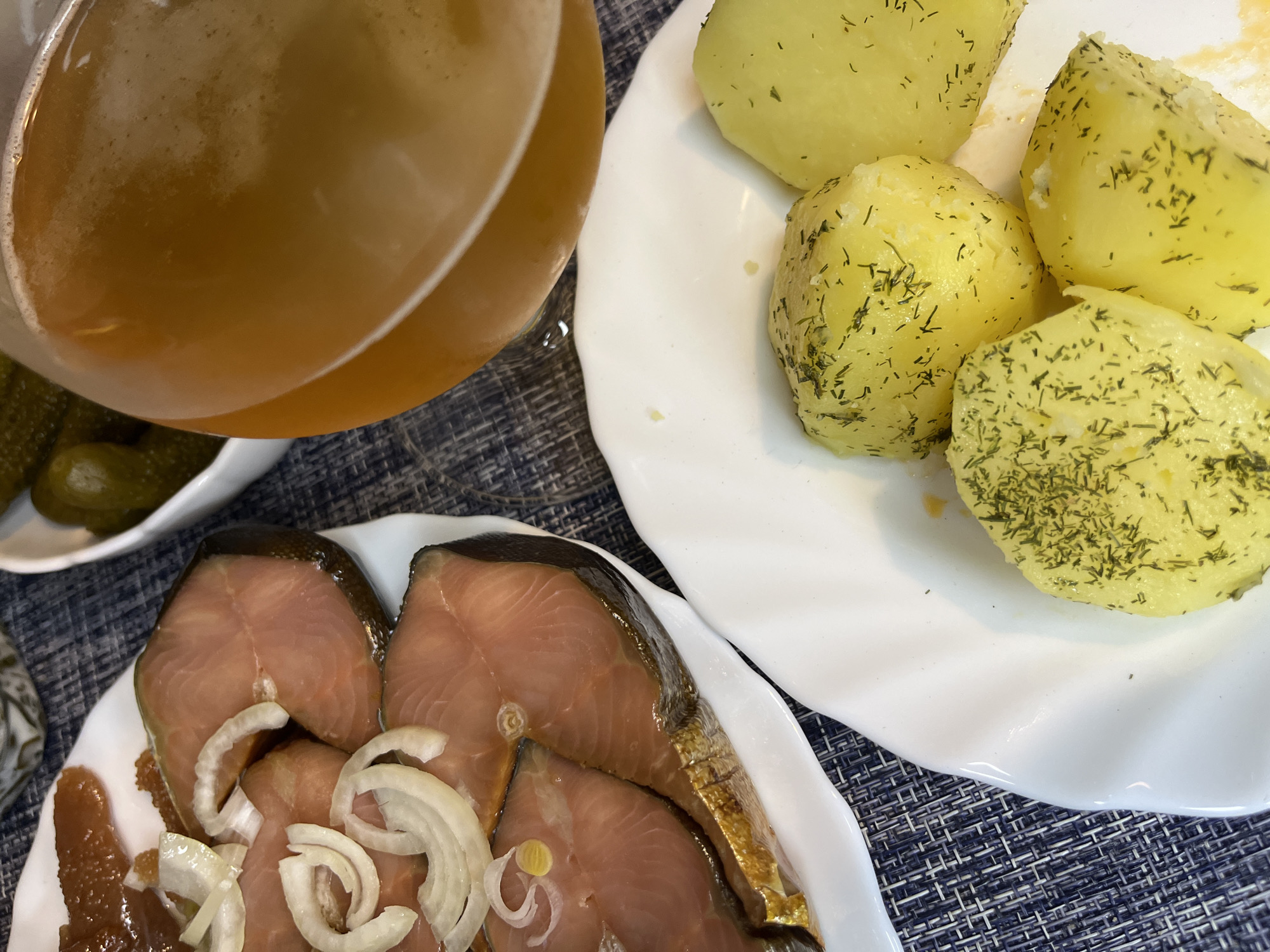 This is a close-up, overhead photograph depicting a meal set on a table. The table surface is covered by a dark blue and white woven placemat.
The meal consists of several dishes. In the upper right section, a white plate holds portions of light yellow, cooked potatoes, which are generously sprinkled with finely chopped green herbs, likely dill. Towards the bottom left, another white plate presents several thick, pinkish slices of what appears to be smoked or cured fish, identifiable by its distinct color and the dark edges of the skin. These fish slices are garnished with translucent rings of white onion and accompanied by a reddish-brown substance, possibly fish roe or a savory spread.
A large glass, filled with an amber-colored, slightly foamy liquid—most likely beer—occupies the upper left side of the frame. Partially visible in the background, to the left of the glass, is a white bowl containing several green pickled cucumbers. The setting appears to be indoors, possibly a home, with artificial lighting, suggesting a meal taking place in the evening. The combination of these dishes is characteristic of traditional Russian cuisine, aligning with the provided location context of Раздолье, Russia. There are no people or visible text in the image.