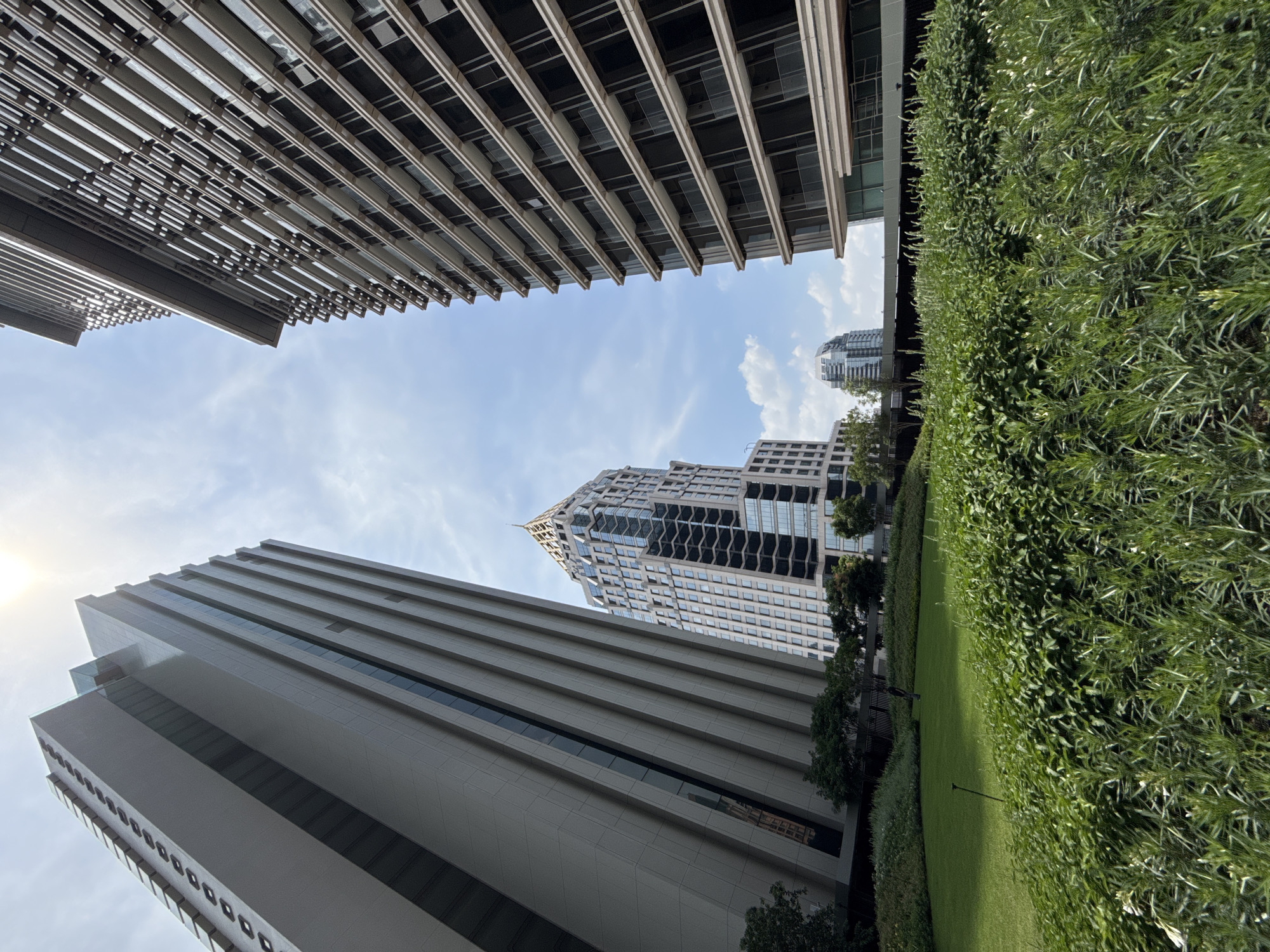 The image, shot from a low angle, depicts a view of tall buildings and greenery in Bangkok, Thailand. In the foreground, a wall of dense green vegetation runs along the right side of the frame. To the left and above, imposing modern buildings dominate the scene. One building features a grid-like exterior with horizontal lines, while another has a smooth, gray facade with dark windows. In the center, a taller building with a distinctive pointed roof can be seen, partially obscured by the surrounding structures. The sky is visible in the spaces between the buildings, revealing a blue sky with scattered clouds. The sun appears to be shining from the left side, casting a subtle glow. The overall perspective emphasizes the scale and density of urban development in Bangkok, contrasted by the presence of green spaces.