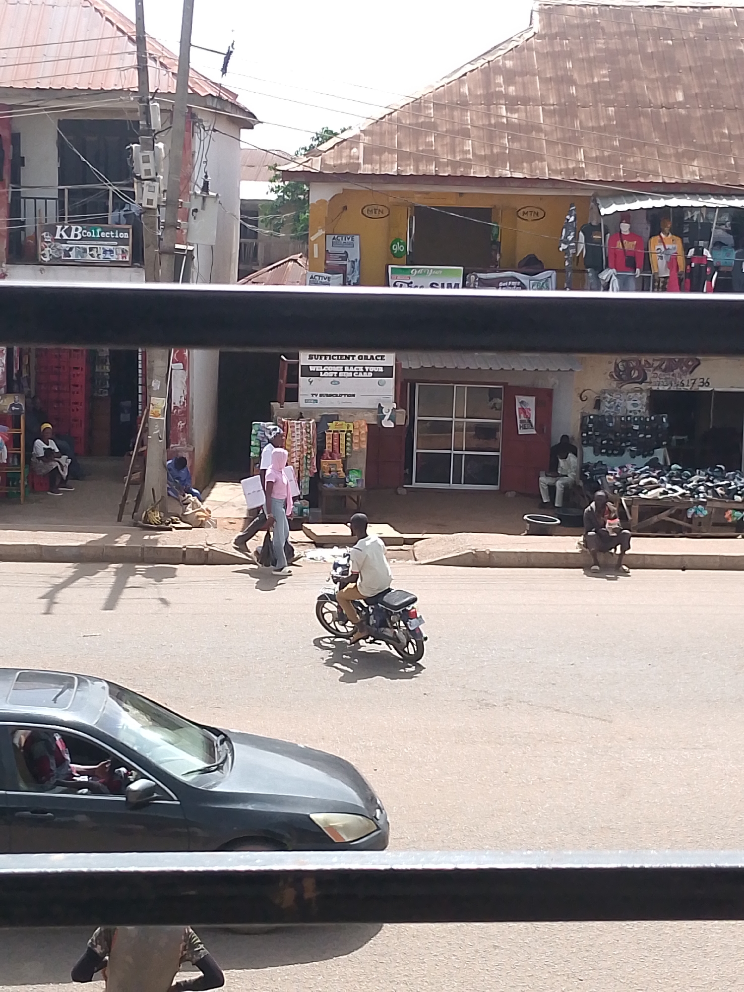 This image depicts a street scene in Bukuru, Nigeria. In the foreground, a dark sedan is parked on the left side of the road, with a person's head visible through the driver's side window. A motorcycle with a rider is traveling on the road to the right of the car.
The background shows a row of shops. The shop on the left has "KBCollection" signage. The shop to its right has "MTN" and "Glo" logos visible. The shop further to the right appears to be a shoe store, with numerous shoes displayed on a wooden rack. People are seen walking on the sidewalk in front of the shops, and some are seated. One person is sitting on the curb, and another person is leaning against a wall. There are also several women sitting on chairs outside one of the shops. The buildings have corrugated metal roofs. The overall environment suggests a busy commercial area.