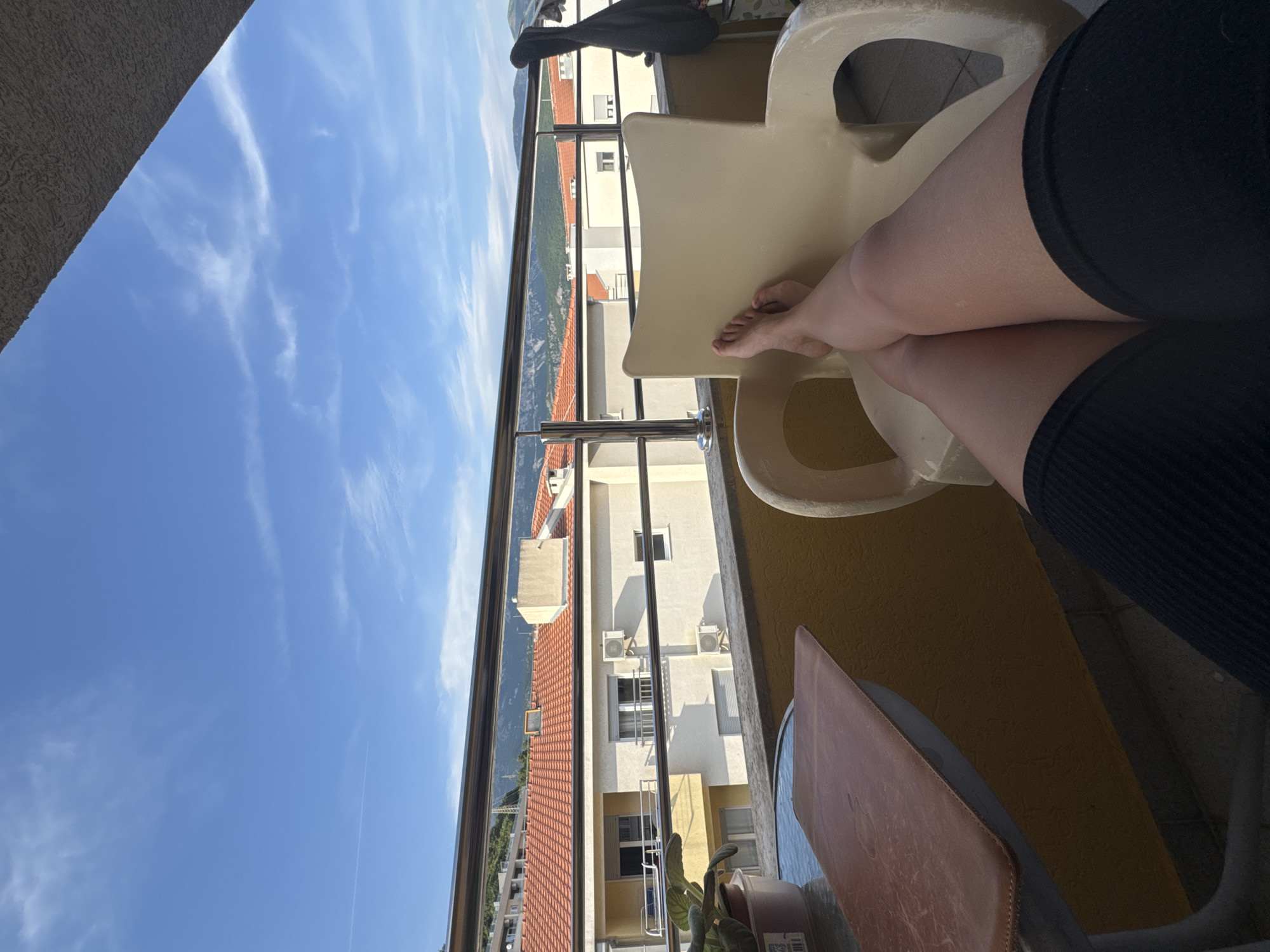 The image depicts a person's legs and feet resting on a beige plastic chair on a balcony. The person is wearing black shorts. In the foreground, a round table with a brown leather-like cover is visible. Behind the railing of the balcony, there are buildings with orange tiled roofs, and in the background, mountains are visible under a blue sky with wispy clouds. The scene appears to be in a city, likely Budva, Montenegro, given the architectural style and the mountainous backdrop common to the region. The lighting suggests it is daytime, possibly late afternoon.