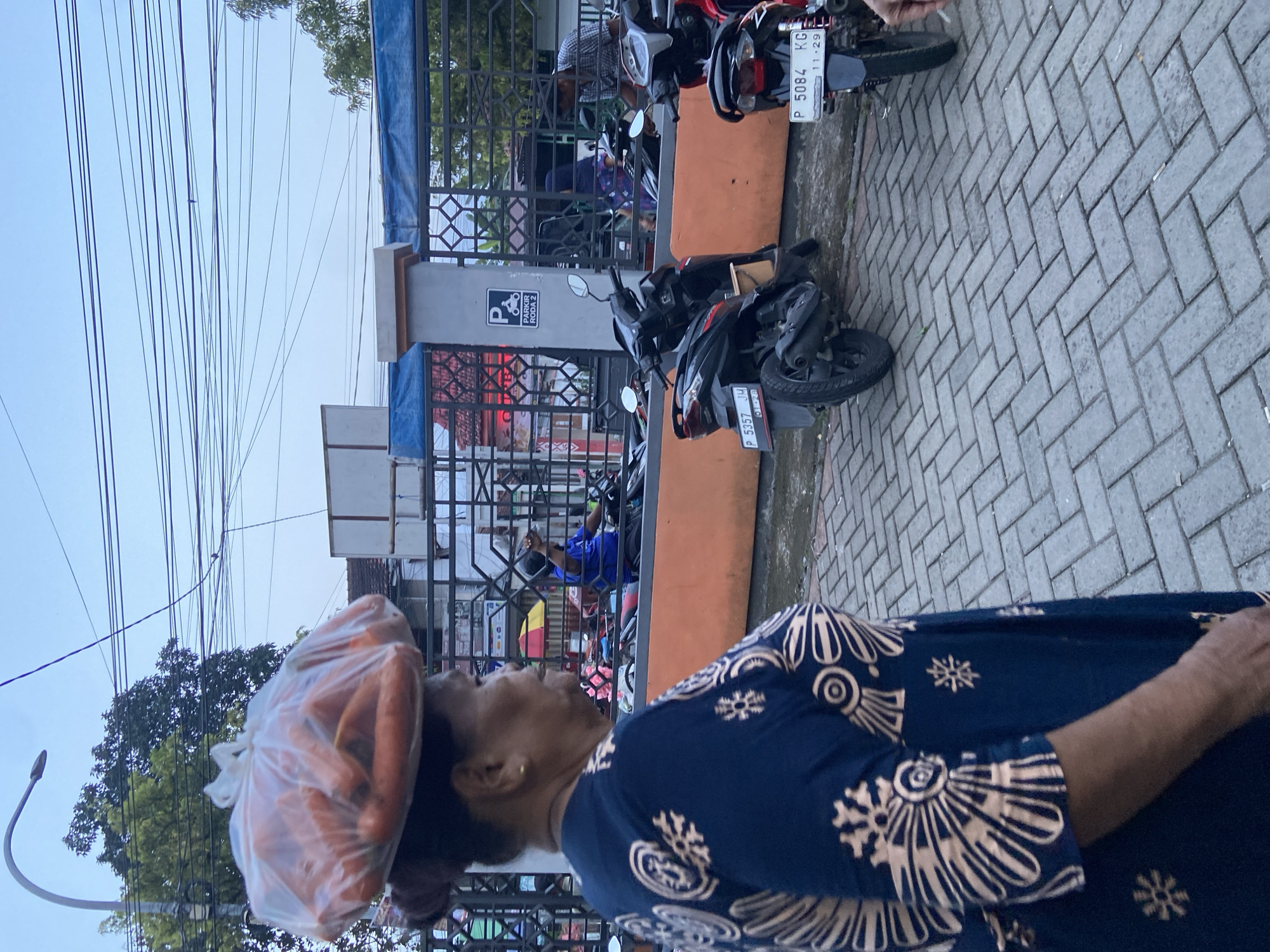 An eye-level shot depicts a woman in Jember, Indonesia, carrying a bag of carrots on her head. The woman is wearing a patterned blue shirt. In the background are motorbikes parked along a building with a sign indicating "PARKER RODA 2". The license plates of the motorbikes read "P 5357 JH" and "P 5084 KG". The ground is paved with interlocking gray bricks. Above, power lines stretch across the sky, partially obscuring a tree.