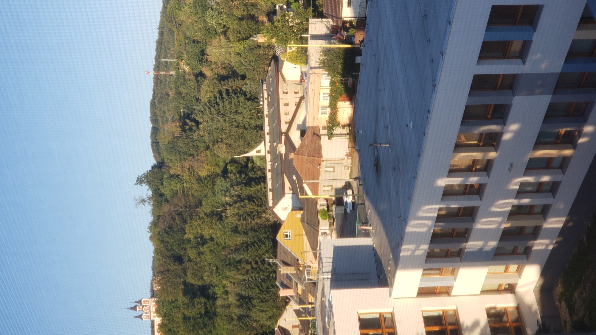 The image captures a view from a building, looking out over a residential area and a tree-covered hill under a clear blue sky. In the foreground, the upper floors of a white and blue modern building with numerous windows are visible, casting long shadows from the sun. Further into the scene, a cluster of houses with red and yellow roofs are nestled amongst trees. A white car is parked on a street between these houses. On the distant hill, a large complex with a distinctive spire-like structure, possibly a church or monastery, is visible on the left. A tall communication tower stands on the right side of the hill. The overall scene suggests a relatively peaceful suburban or semi-urban environment in Prešov, Slovakia, likely captured during a sunny day.