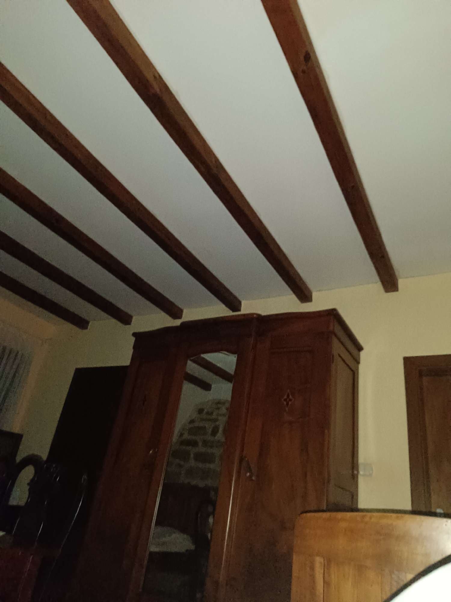 The image shows a bedroom interior with a white ceiling and exposed wooden beams. A large, dark wood wardrobe with intricate detailing is positioned prominently in the center. To the left of the wardrobe, a dark wooden chair with a curved back is partially visible. A section of a stone wall is visible through the open doors of the wardrobe. To the right of the wardrobe, another wooden door is visible. The overall ambiance suggests a traditional or rustic decor. The location is described as Unknown, Unknown.