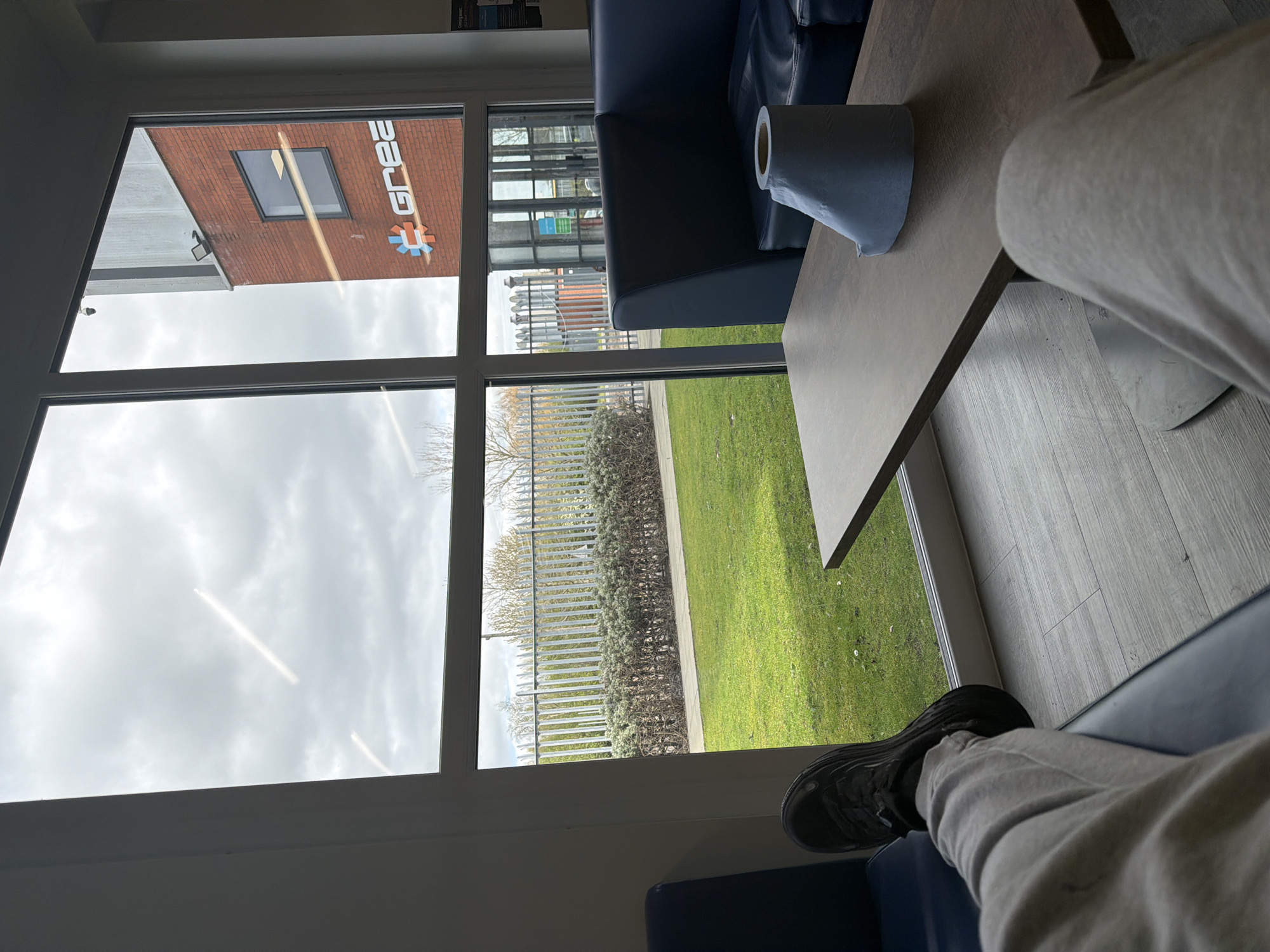 The image, taken from an indoor perspective, looks out through a large window divided by white frames. The view encompasses both interior elements and an outdoor scene. Indoors, the lower half of a person's legs and feet are visible, clad in grey sweatpants and black sneakers, suggesting they are seated. A blue sofa is partially seen, and a light-colored rectangular table occupies the right side of the frame, holding a roll of paper towels.
Outside the window, a prominent red brick building features a large white sign with the word "GREA" and a distinctive red and blue cog-like logo. A dark, rectangular window is also visible on the brick facade. The outdoor landscape includes a vibrant green grassy area, a metallic fence, and a line of dense bushes. Bare trees are discernible in the distance under an overcast but bright sky, indicating daytime. The scene suggests a commercial or industrial setting, possibly a waiting area or office lounge, in Connah's Quay, United Kingdom. No specific activity is ongoing apart from the person relaxing indoors.