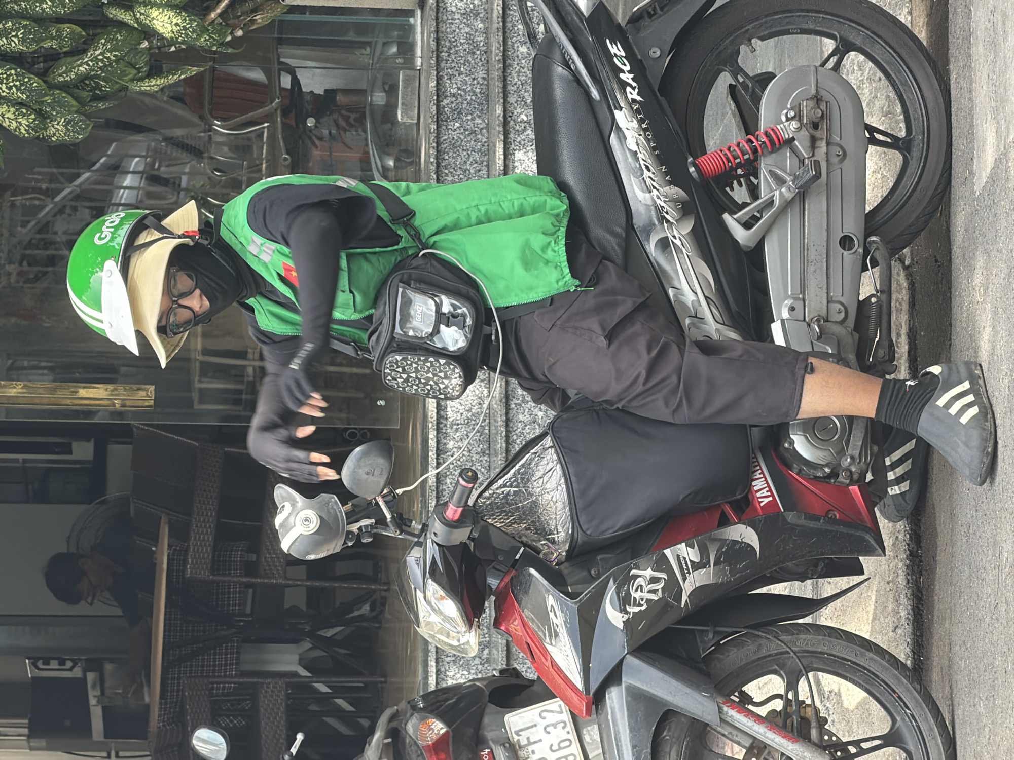 A person wearing a green Grab helmet, a tan hat, glasses, a black face mask, a black long-sleeved shirt, a green vest, and black pants is sitting on a black and red motorcycle. The motorcycle has a license plate that reads "F1 96.32". The person is wearing black gloves and black slip-on shoes with white stripes. A black bag with a clear front pocket and the word "GENZ" is attached to the person's waist. The motorcycle is parked on a paved surface in front of a building with large glass windows and an awning. In the background, a person is seated at a table inside the building. The scene appears to be on a street in Nha Trang, Vietnam, indicated by the "Grab" branding and the general streetscape.