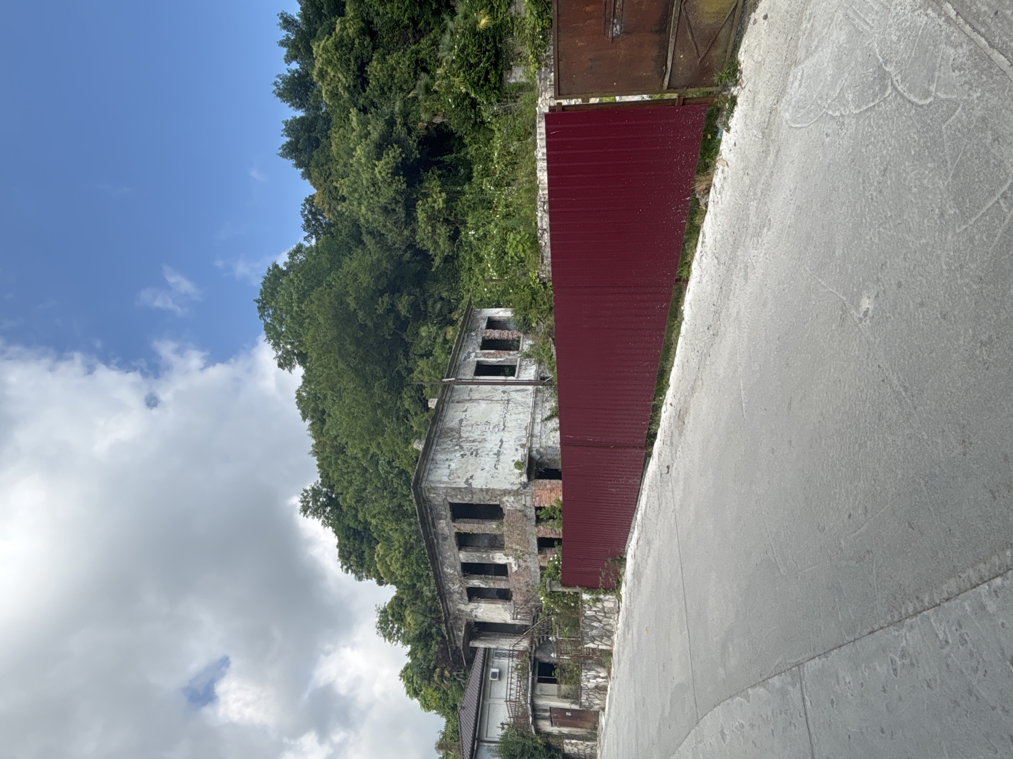 This is a vertical, eye-level, full shot of an abandoned building nestled in a lush, green landscape. The building appears to be several stories tall and made of concrete and brick. It is in a state of disrepair, with gaping windows and weathered walls. The surrounding vegetation is thick and overgrown, with trees and foliage encroaching on the structure. A tall, dark red corrugated metal fence runs along the side of the building, separating it from a concrete path or road in the foreground. Behind the building, a dense forest climbs a steep incline under a partly cloudy sky. The sky is a bright blue with white, fluffy clouds. The lighting suggests it is daytime, possibly late morning or early afternoon, given the brightness of the sky. The scene evokes a sense of abandonment and decay, with nature reclaiming the man-made structure. No people are visible in the frame. There is no visible text in the image.