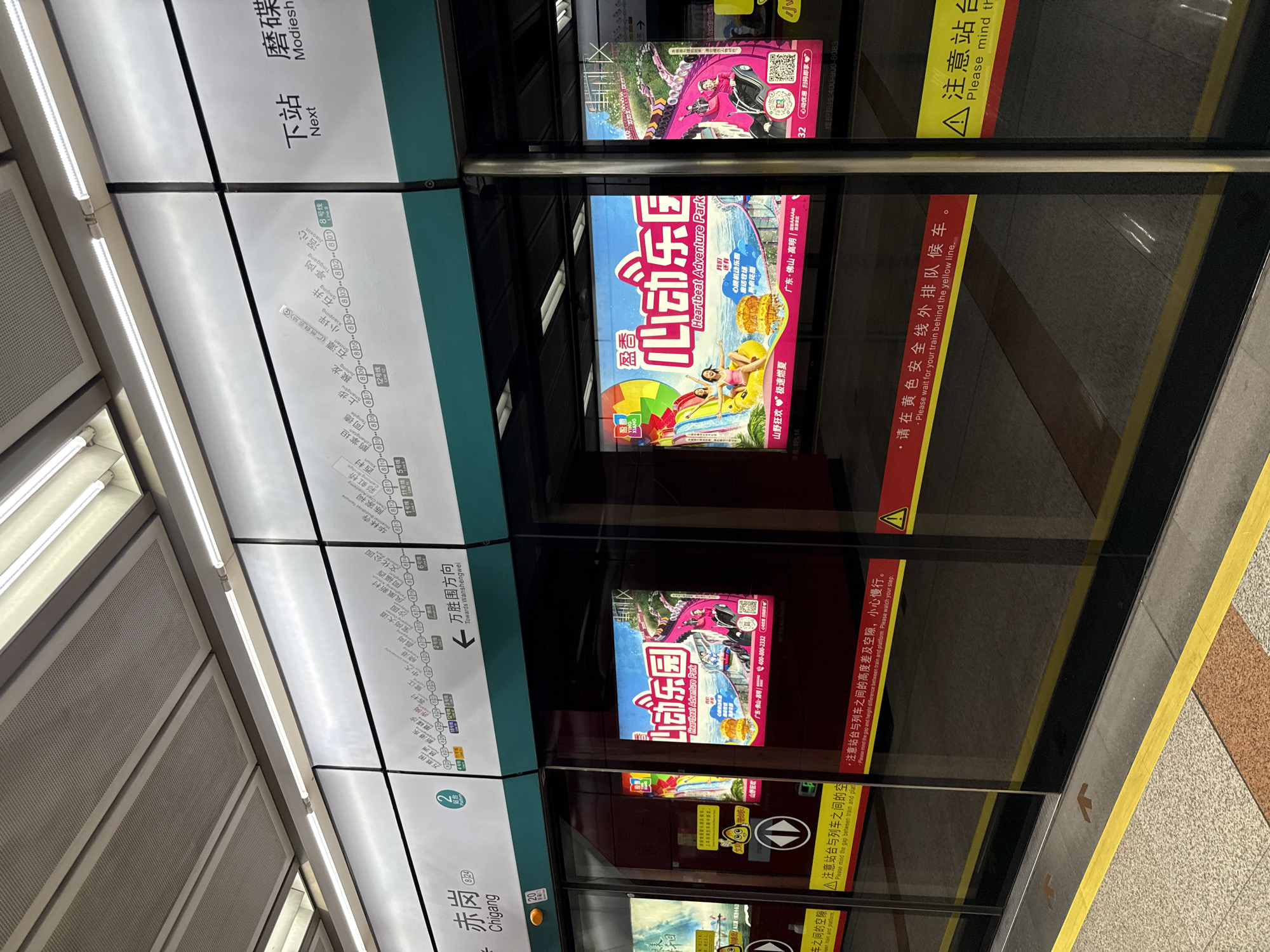 The image captures a subway platform in Haizhu District, China. The platform features glass safety doors that partially obscure the view of the tracks and advertisements displayed on the doors themselves. Above the platform, a digital map of the subway lines is visible, showing station names and line numbers. Prominently displayed on the platform doors are two brightly colored advertisements for "Heartbeat Adventure Park." The advertisements feature imagery of water slides, amusement park rides, and happy people enjoying themselves. The station itself appears clean and modern, with white walls and ceiling lighting. A yellow safety line is visible on the platform floor, indicating where passengers should stand while waiting for the train. The overall scene is typical of a busy urban subway station.