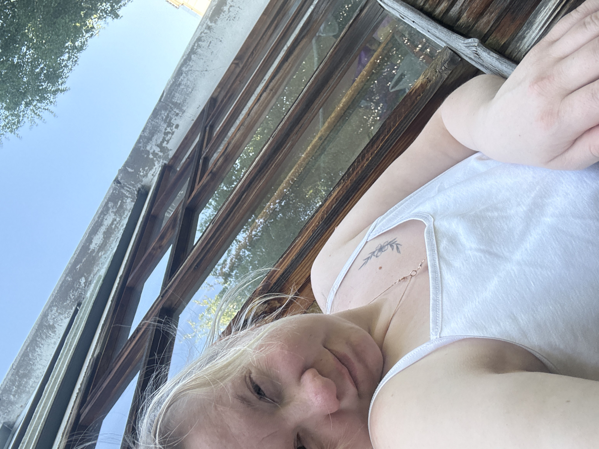 A woman wearing a white tank top is posing for a selfie. She is under a dilapidated wooden structure with peeling paint. The scene is outdoors, with the sky and foliage visible in the background. The location is Chaikovsky, Russia.