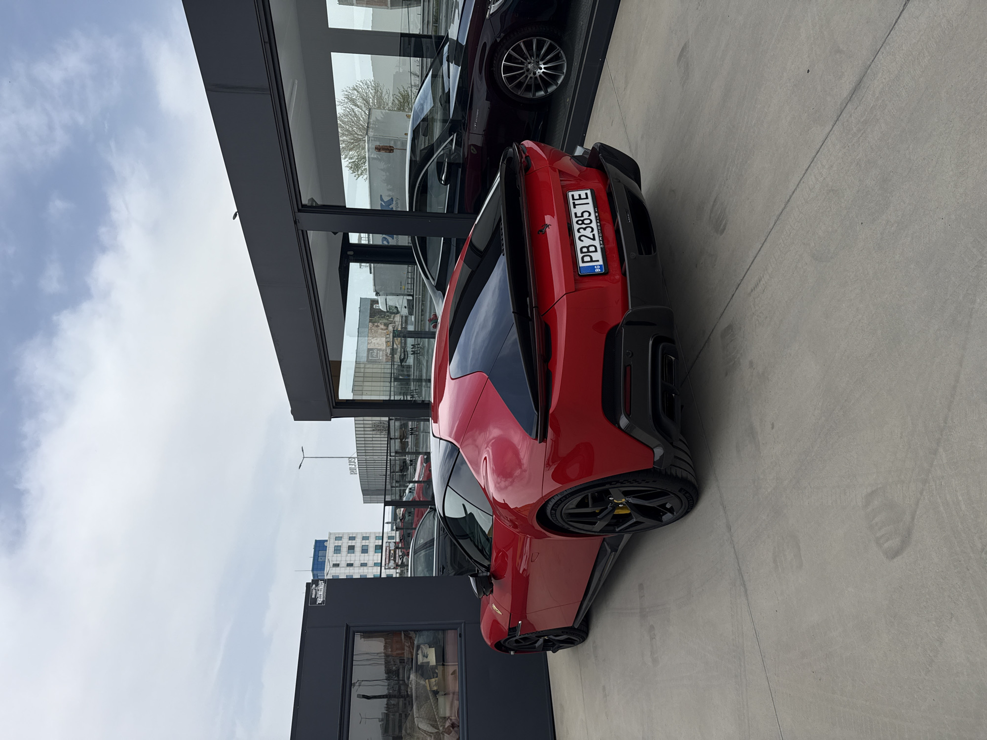 A red Ferrari coupe, displaying a Bulgarian license plate "PB 2385 TE," is parked in front of a dealership. Other cars are visible through the dealership windows. The sky is partly cloudy. The location is in Plovdiv, Bulgaria.