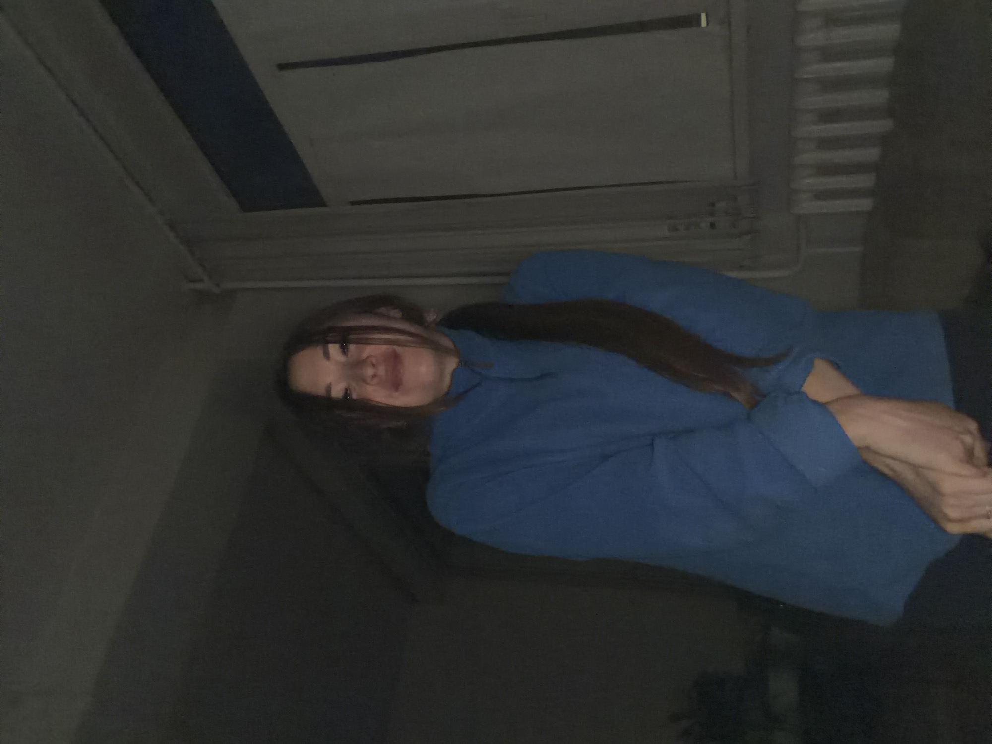 The image shows a young woman with long, dark hair, wearing a blue sweatshirt. She is positioned in a way that her face is tilted upwards and her body is angled towards the viewer. Her arms are crossed in front of her. The background appears to be indoors, with a ceiling that has some structural elements and a blue stripe. There are also some horizontal lines suggesting ventilation or shelving. The lighting is dim, creating shadows and highlighting parts of her face and arms. The overall mood is casual and personal, possibly a selfie. The provided location context is Япрыково, Russia. No text is visible in the image.