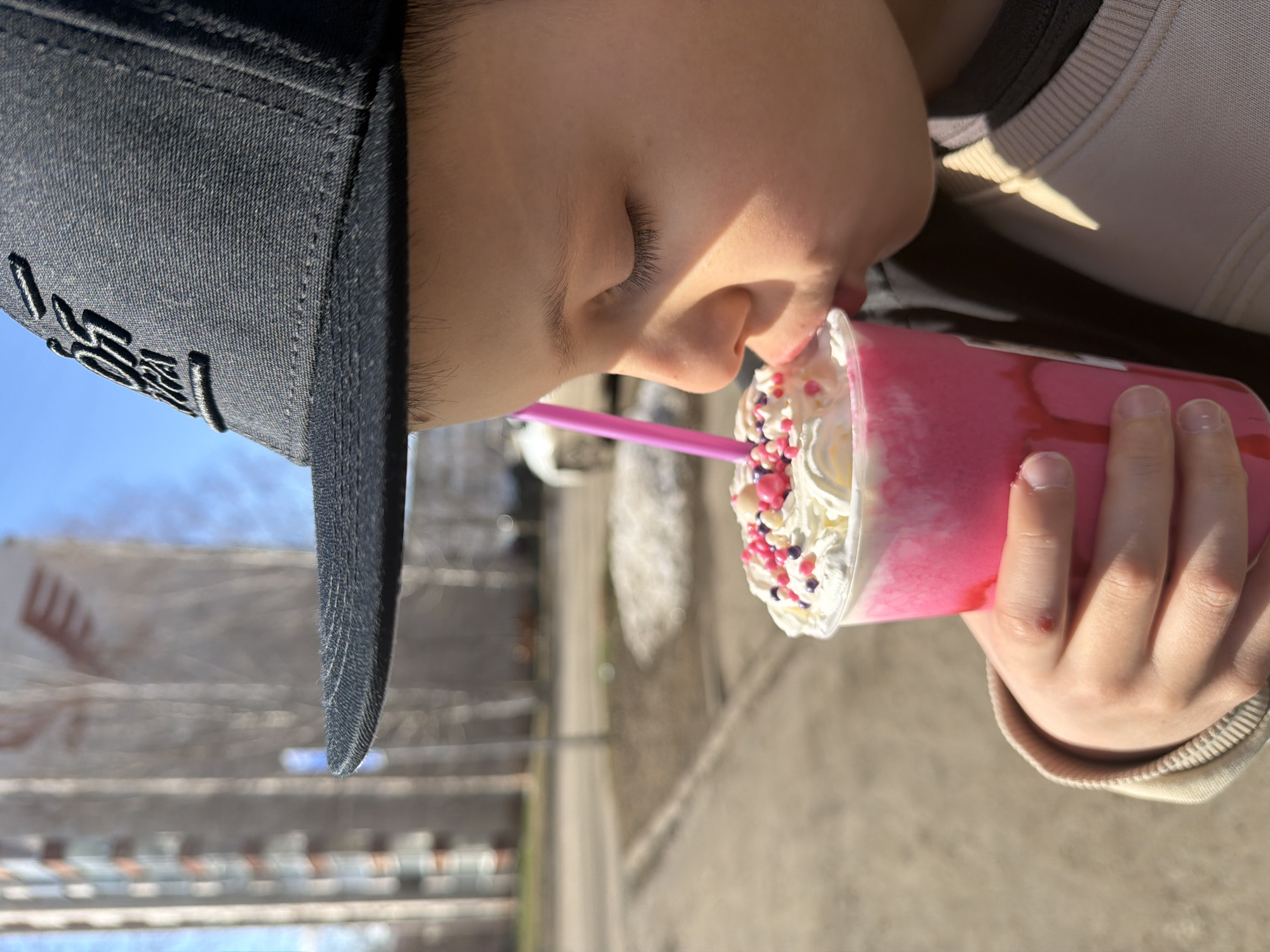 A child wearing a black baseball cap is drinking a pink milkshake with whipped cream and sprinkles through a pink straw. The child is wearing a cream-colored shirt with a black collar. The background shows a street and buildings, possibly located in a city, based on the built environment. The location is Votkinsk, Russia.