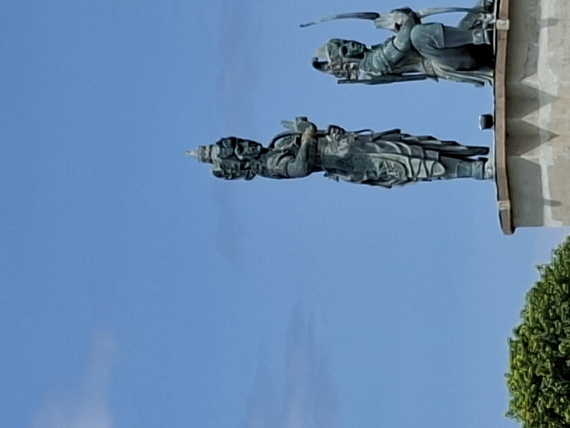 A photograph shows two statue figures on a stone base, set against a clear blue sky. The lower figure, likely a warrior or deity, is depicted in a detailed metallic material. The statue is crowned and appears to be in a pose that suggests prayer or an offering of peace. Above and slightly behind, there is another statue figure, possibly a winged being or guardian, with similar detailing. The presence of green foliage at the bottom right corner suggests that the statues are likely located in an outdoor or garden area. These sculptures are part of Garuda Wisnu Kencana Cultural Park, located in Kuta Selatan, Bali, Indonesia.