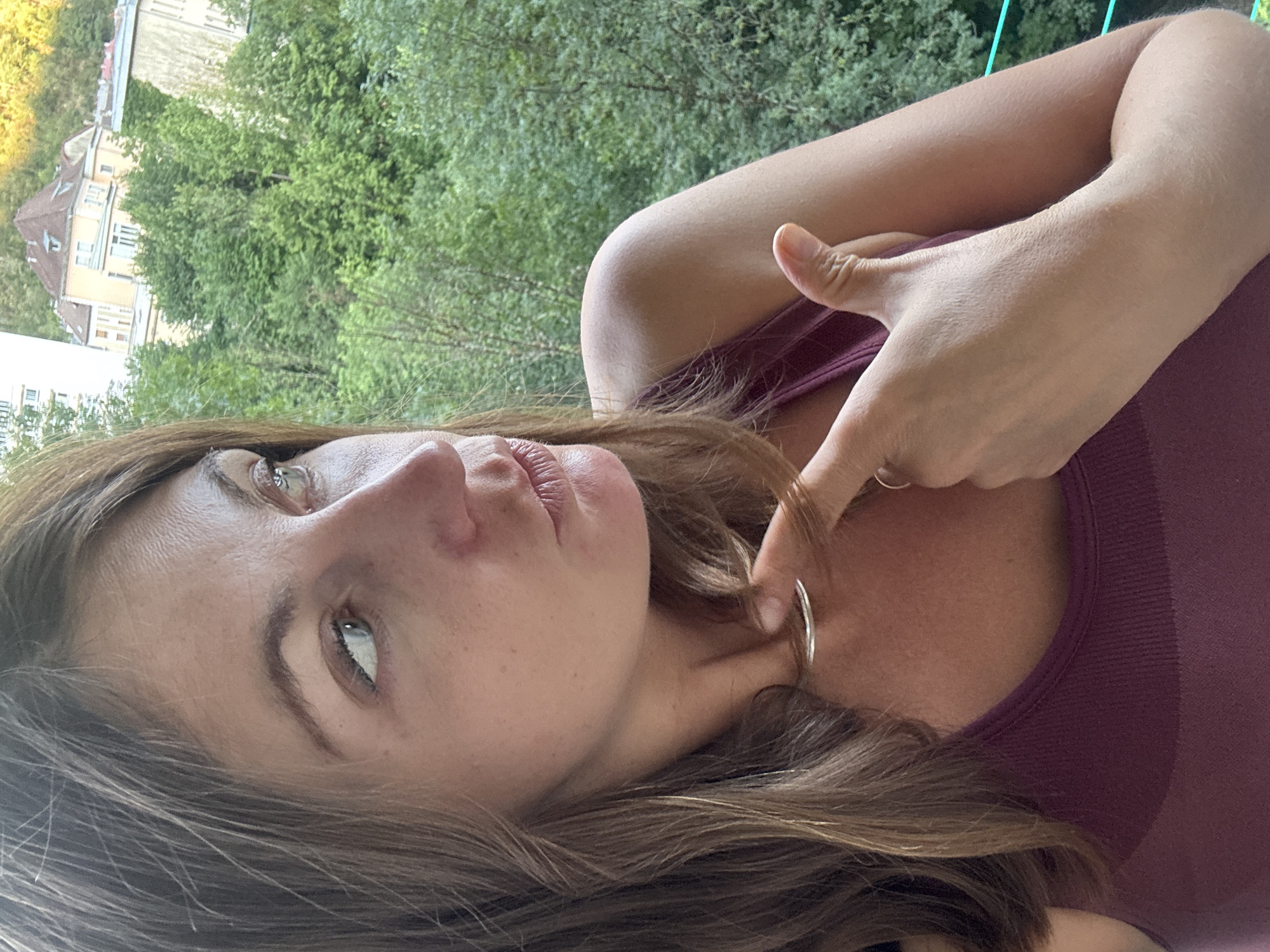 The image captures a close-up of a woman's face and upper body, taken from a high vantage point that overlooks a cityscape with lush green trees and buildings. The woman, with shoulder-length brown hair and light blue eyes, is looking to her right, with her right hand touching her neck, where a thin silver necklace is visible. She is wearing a dark reddish-purple tank top. In the background, a multi-story building with a reddish-brown roof and several windows is prominent, nestled amongst dense green foliage. The urban setting suggests a location within a city, and given the previous information, it is identified as Saint-Claude, France. The lighting indicates it is daytime, with a warm glow suggesting either morning or late afternoon sun.