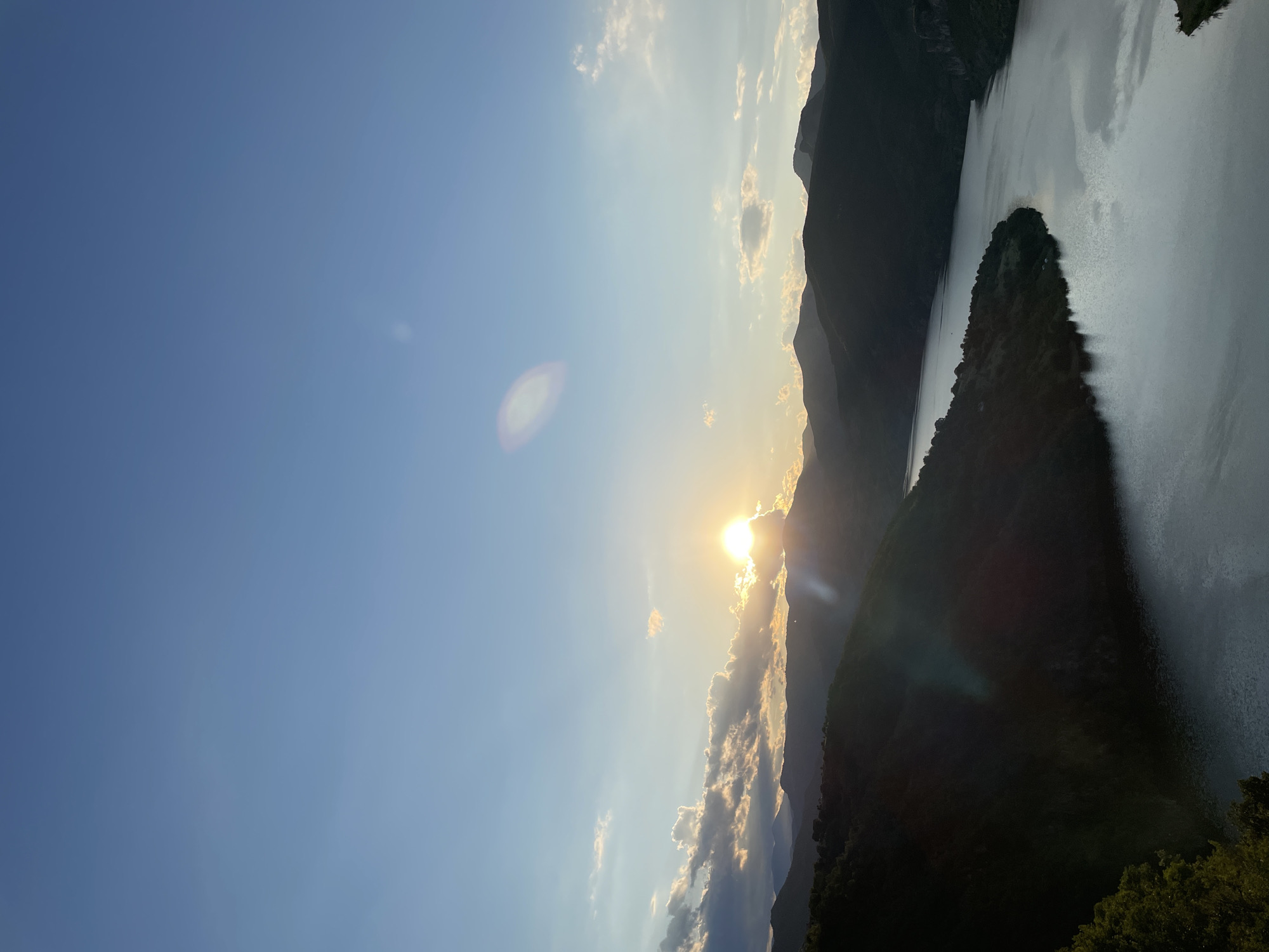 The image shows a sunset over a mountain landscape with a river. The sun is low on the horizon, casting a warm glow on the clouds and the mountains. The river flows through a valley, reflecting the colors of the sky. The scene is serene and peaceful, with a sense of natural beauty. The location is identified as Borovitsa, Bulgaria. There are no people or actions depicted in the image. The environment is natural, with mountains, a river, and a sky with clouds.