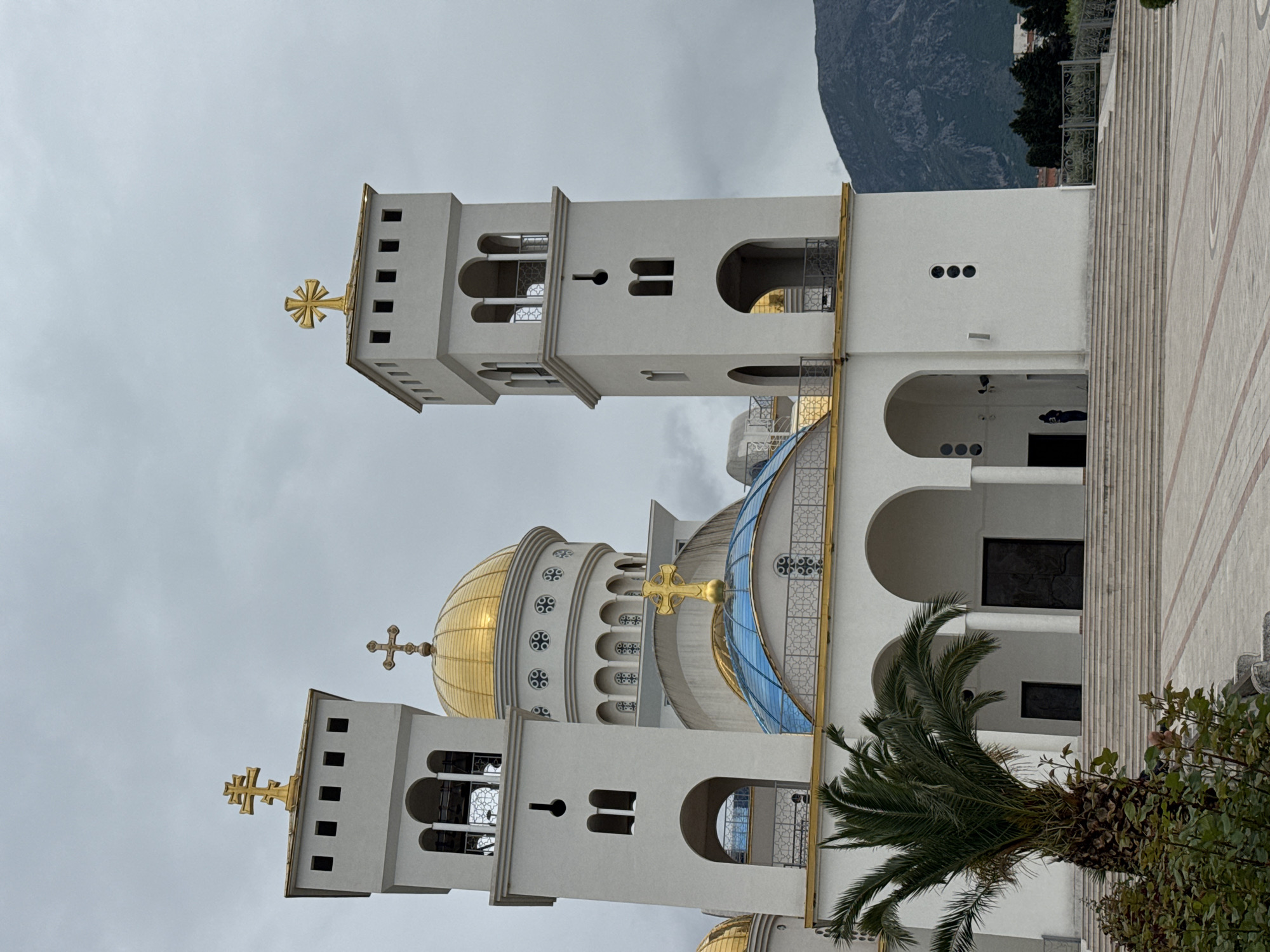 The image displays a grand, white Eastern Orthodox cathedral set against an overcast sky and a mountainous backdrop. The main structure features multiple golden domes, including a large central one and several smaller ones, all surmounted by distinctive golden Orthodox crosses with slanted lower bars. A prominent blue-tinted glass dome is also visible as part of the complex.
Two tall, rectangular bell towers, also white, flank the central dome, each topped with a golden cross. The towers exhibit arched belfry openings and smaller square windows. The cathedral's architecture is characterized by its stark white walls, intricate gold-colored railings and grilles, and decorative patterns such as circles adorning the base of the domes. Arched entrances and windows are scattered across the building's facade.
In the foreground and to the side, a vibrant green palm tree and other leafy vegetation are visible. The ground area leading up to the cathedral is paved with light-colored, patterned tiles and steps. A large, dark, forested mountain rises in the background, providing a dramatic setting.
The weather is overcast, contributing to a muted light quality. No people or specific events are depicted, and no visible text is present. The scene conveys a sense of architectural splendor and spiritual significance in a natural, mountainous environment.