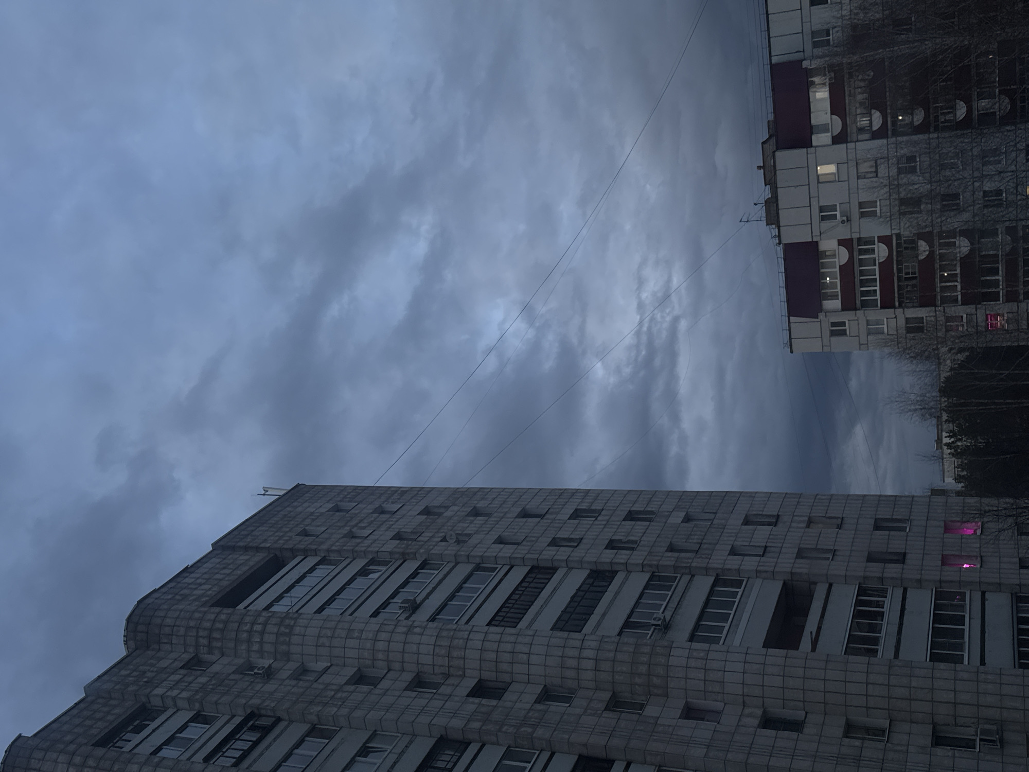 This image, presented rotated 90 degrees clockwise, captures an urban scene in Chaikovsky, Russia, under a heavily overcast and gloomy sky. The sky is filled with thick, grey-blue clouds, suggesting either dusk or a deeply shadowed daytime.
Two multi-story buildings dominate the landscape. The most prominent structure, occupying the lower left and center (when viewed correctly), features a distinctive, heavily textured facade composed of square or rectangular panels. This building, likely an apartment block or office building, exhibits numerous windows, some of which are open or fitted with external air conditioning units. A few windows on this building emit a bright pink or purple light, providing a striking color contrast against the otherwise monochromatic palette.
To the upper right (when viewed correctly), another residential building is partially visible. This one presents a more conventional appearance with balconies, lighter grey walls, and sections of dark red or purple on its roof. Several windows in this building are illuminated, indicating the presence of occupants. Bare tree branches, suggesting a late autumn or winter season, can be seen adjacent to this structure.
Thin power lines or utility cables stretch across the cloudy expanse. The overall mood is one of quiet urban utility, potentially evoking a sense of everyday life in a post-Soviet environment. No discernible activity or event is taking place, and there is no visible text within the frame. The dim lighting reinforces the impression of twilight or an overcast day.
