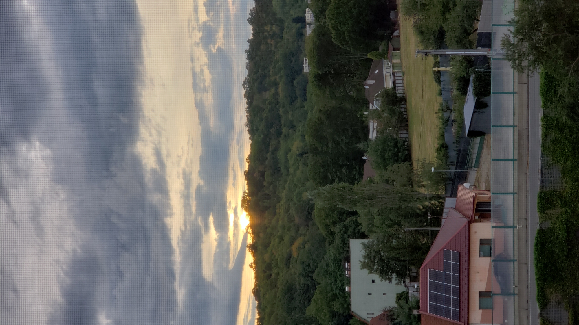 The image depicts a sunset over a wooded hillside in Prešov, Slovakia, as viewed through a window screen. In the foreground, a house with a red roof and solar panels is visible, along with a road and a grassy area. The hillside is covered with dense green trees, and the sky is filled with dramatic clouds, with the sun peeking through on the horizon. The scene appears to be a residential area on the outskirts of the city.