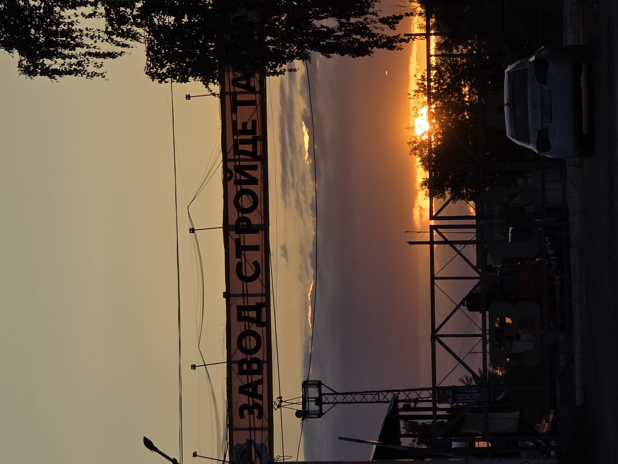 The image presents a striking sunset scene over an industrial or urban landscape in Chaikovsky, Russia. The time of day is clearly evening, with the sun a brilliant, fiery orange orb low on the right side of the horizon, casting an intense glow across the sky. The sky transitions from bright orange and yellow near the sun to softer purples and grays, with scattered clouds illuminated from below, highlighting their edges with warm light.
The foreground elements are largely in stark silhouette against this vibrant backdrop. A large banner or billboard stretches horizontally across the mid-left portion of the frame, displaying Cyrillic text. The visible text reads "ЗАВОД СТРОЙДЕТА...", which translates to "Factory of Building Parts" or "Construction Part Factory." Below and around this sign, various metal structures, possibly scaffolding, industrial frames, or a part of a crane, are silhouetted.
Silhouetted trees and dense foliage frame the upper left and upper right parts of the scene. On the far right, a light-colored sedan car (possibly white or silver) is parked, also in silhouette, with its rear visible. Overhead power lines or cables are strung across the sky, and a street light is visible among the foreground structures. The scene is static, capturing a peaceful yet dramatic moment at the end of the day, contrasting the natural beauty of the sunset with the man-made industrial environment.