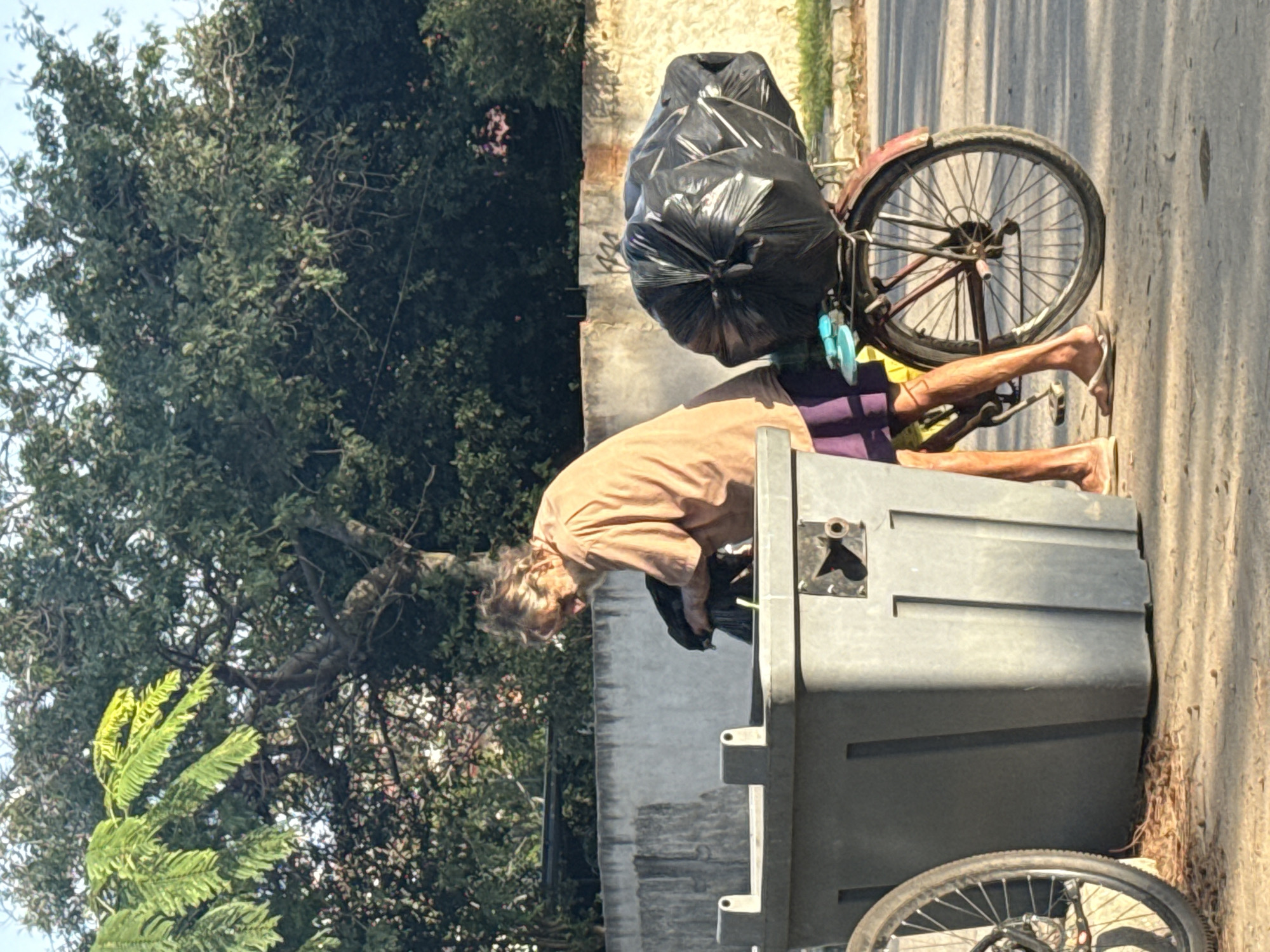 The image depicts a street scene in Iguaba Grande, Brazil, focusing on a man in his 50s or 60s with a beard and disheveled hair, wearing a light-colored t-shirt and shorts. He is standing next to a large, grey trash receptacle, appearing to be interacting with its contents. A bicycle with a large black garbage bag strapped to its rear rack is positioned to the man's right. The background shows trees and a wall, suggesting an outdoor urban environment. The lighting indicates it is daytime, with the sun casting shadows. The overall atmosphere is one of daily life in the city, with an emphasis on resourcefulness and the urban landscape.