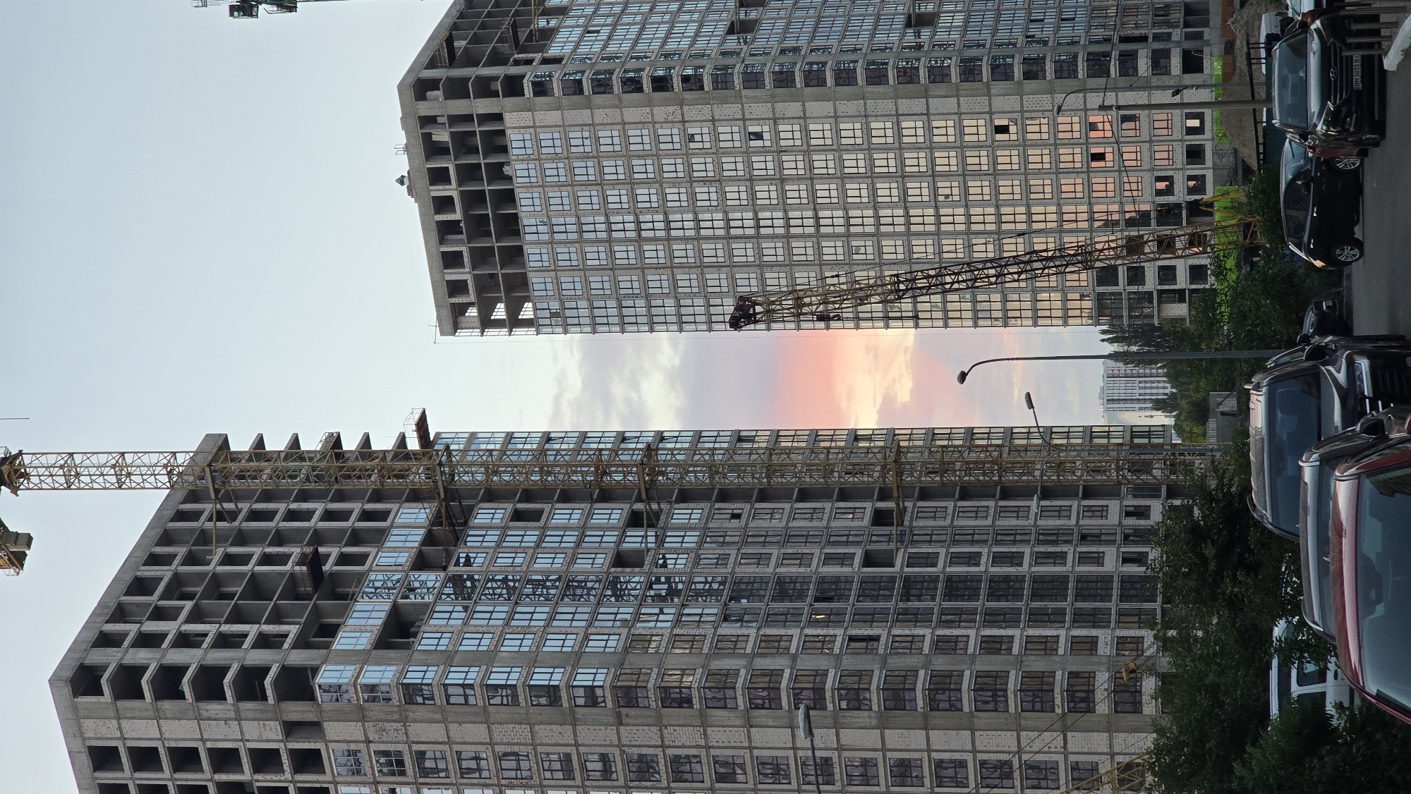 In Kyiv, Ukraine, two unfinished high-rise buildings dominate the scene. A tall construction crane extends upwards between the buildings, its yellow lattice structure stark against the pale sky. The buildings themselves are concrete shells, with the lower floors of one structure already fitted with rows of reflective windows, mirroring the soft pink and orange hues of the sunset. Trees and dense foliage line the foreground, partially obscuring a street where several cars are parked. The overall impression is one of ongoing urban development in a city setting.