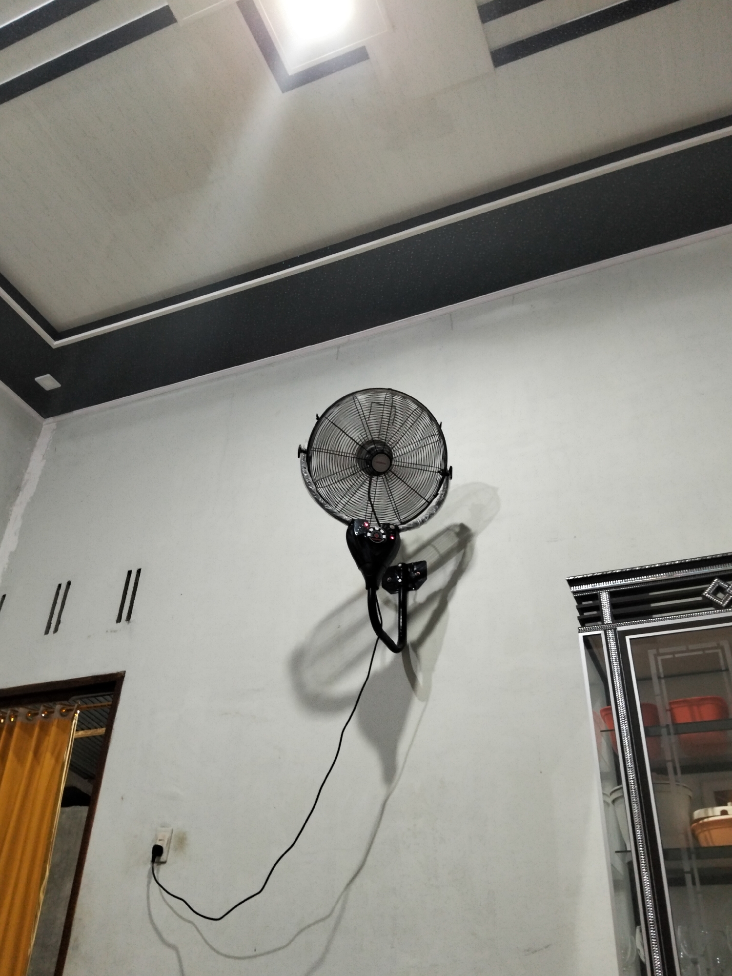 In the city of Cottrueng, Indonesia, a wall-mounted electric fan is depicted hanging on a white wall. The fan is black and circular with visible blades and a metallic grill. A black cord extends from the fan, plugged into a white electrical outlet on the wall below. The fan casts a prominent shadow on the wall. Above the wall, a white ceiling is visible with dark grey decorative trim and a square light fixture. To the left, a doorway with a yellow curtain is partially visible. To the right, a cabinet with glass doors and a decorative frame is present, containing shelves with various orange and white containers. The overall scene appears to be an interior shot of a room.